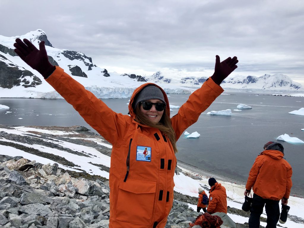 raveler in an orange parka smiling with arms outstretched on Cuverville Island during an Antarctica cruise, with snowy mountains, icebergs, and the ocean behind her.