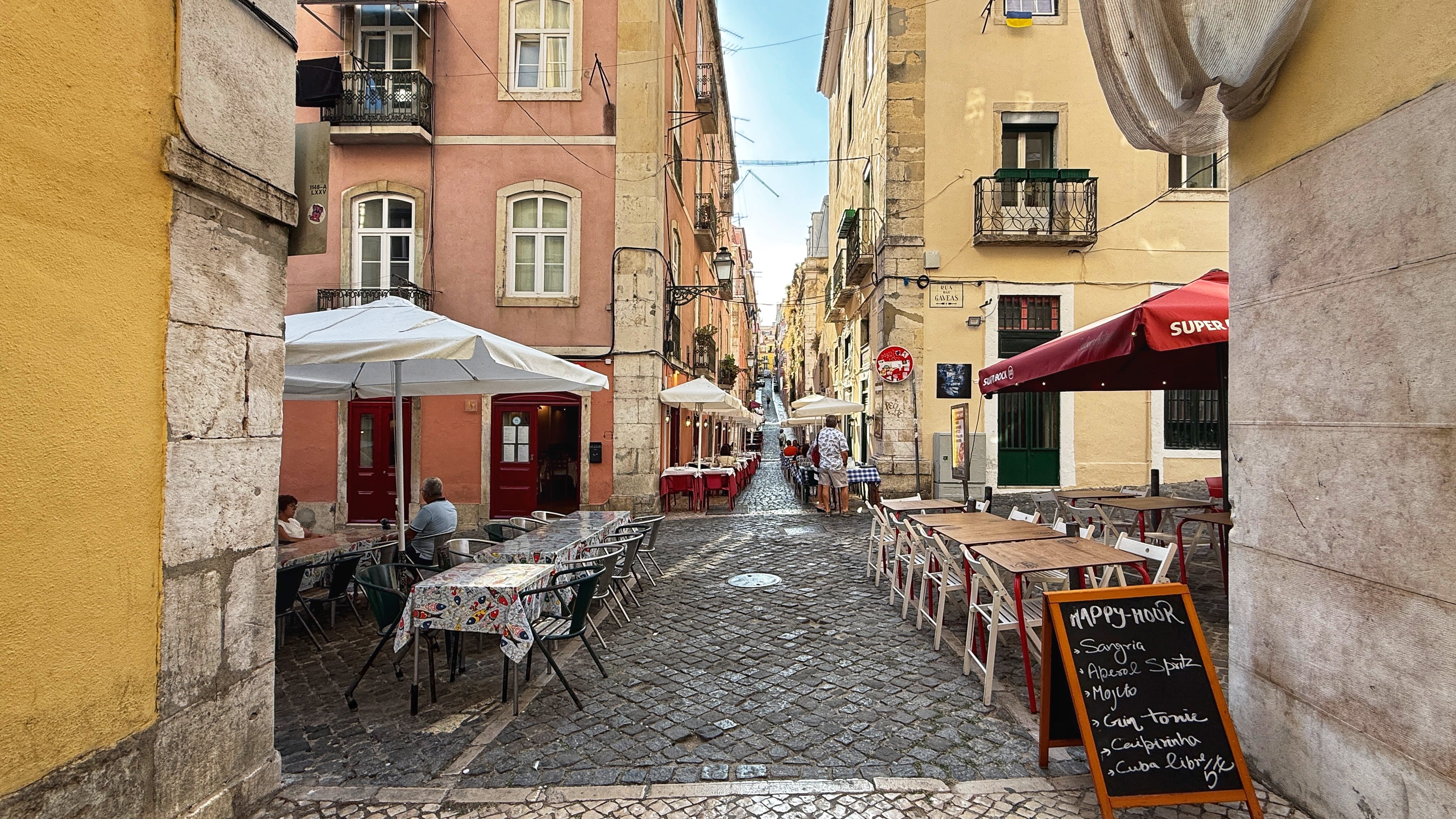 Outdoor café tables lining a cobblestone alley in Lisbon, with pastel-colored buildings, umbrellas, and a “Happy Hour” chalkboard sign in the foreground —5 Days in Lisbon