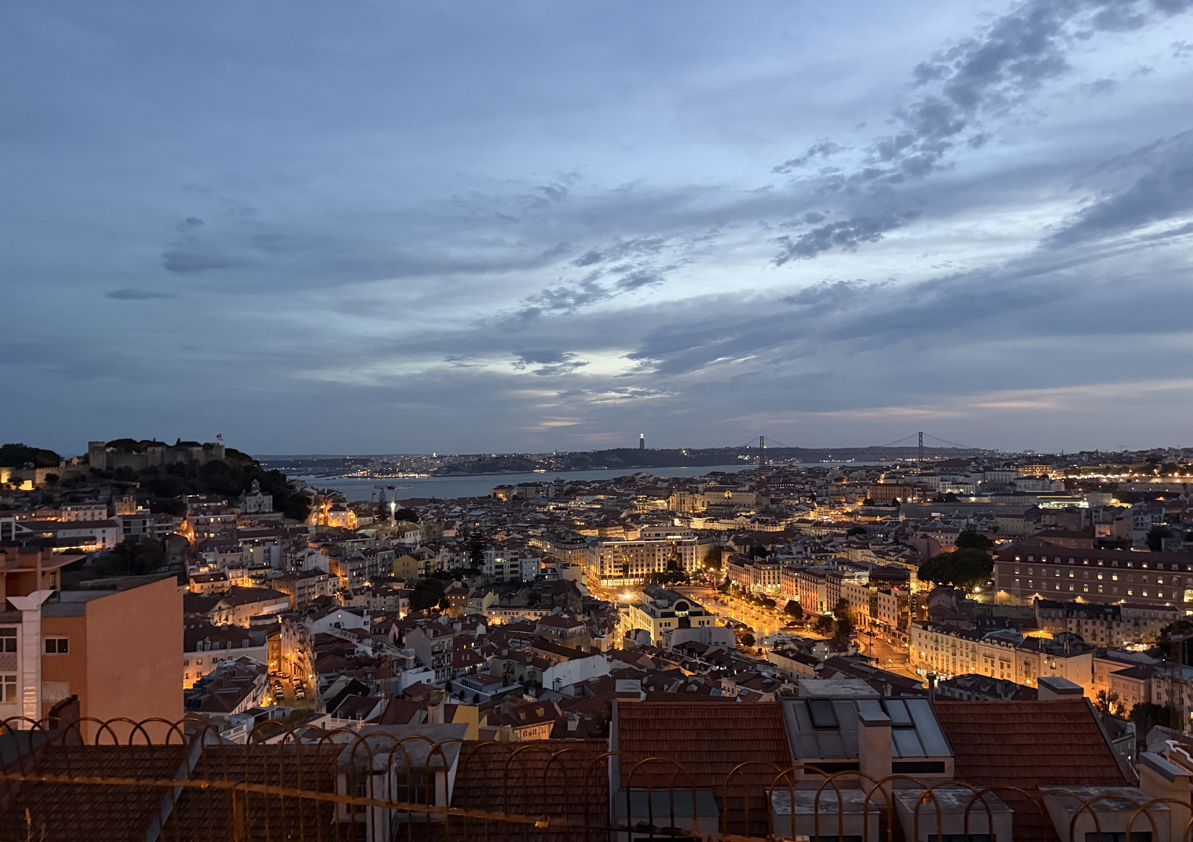 Evening view of Lisbon from a high vantage point during a sidecar tour, with Castelo de São Jorge on the left, the Tagus River in the distance, and the 25 de Abril Bridge under a dramatic sky—5 Days in Lisbon