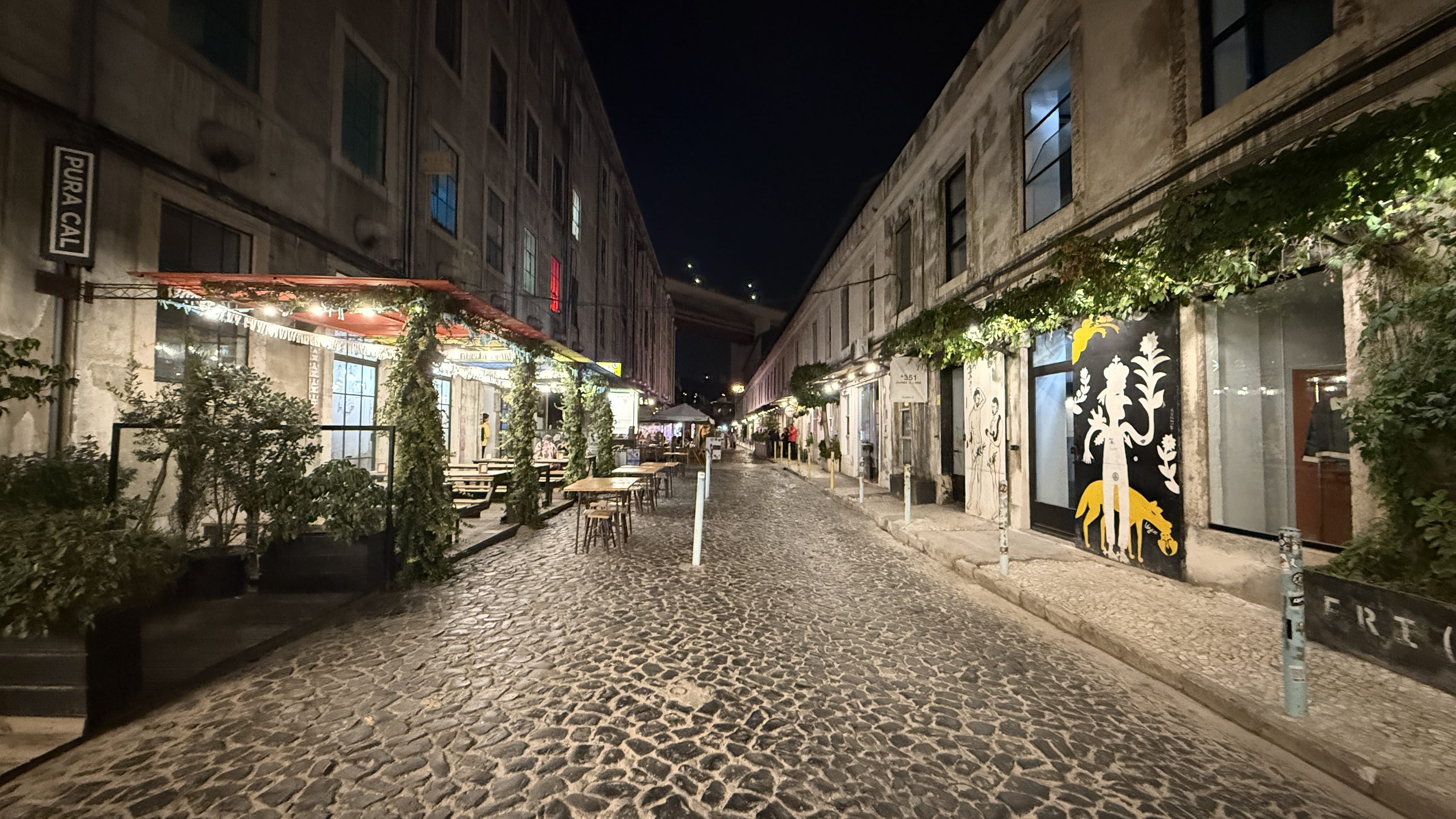 Night view of a cobblestone street in Lisbon’s LX Factory district, lined with old industrial buildings converted into cafés, bars, and shops, with outdoor tables and murals on the walls.