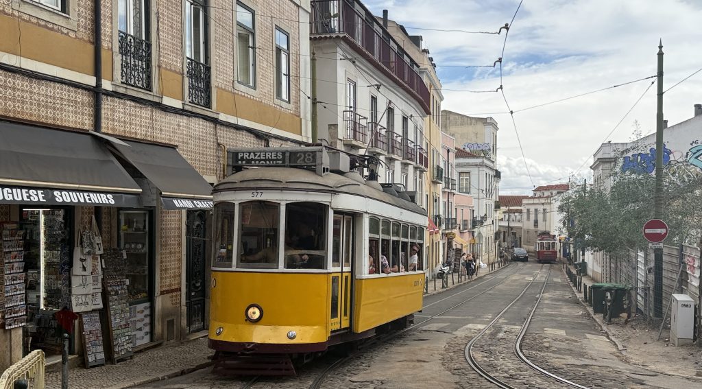 Yellow Tram 28 traveling through a hilly Lisbon street lined with tiled buildings and shops.