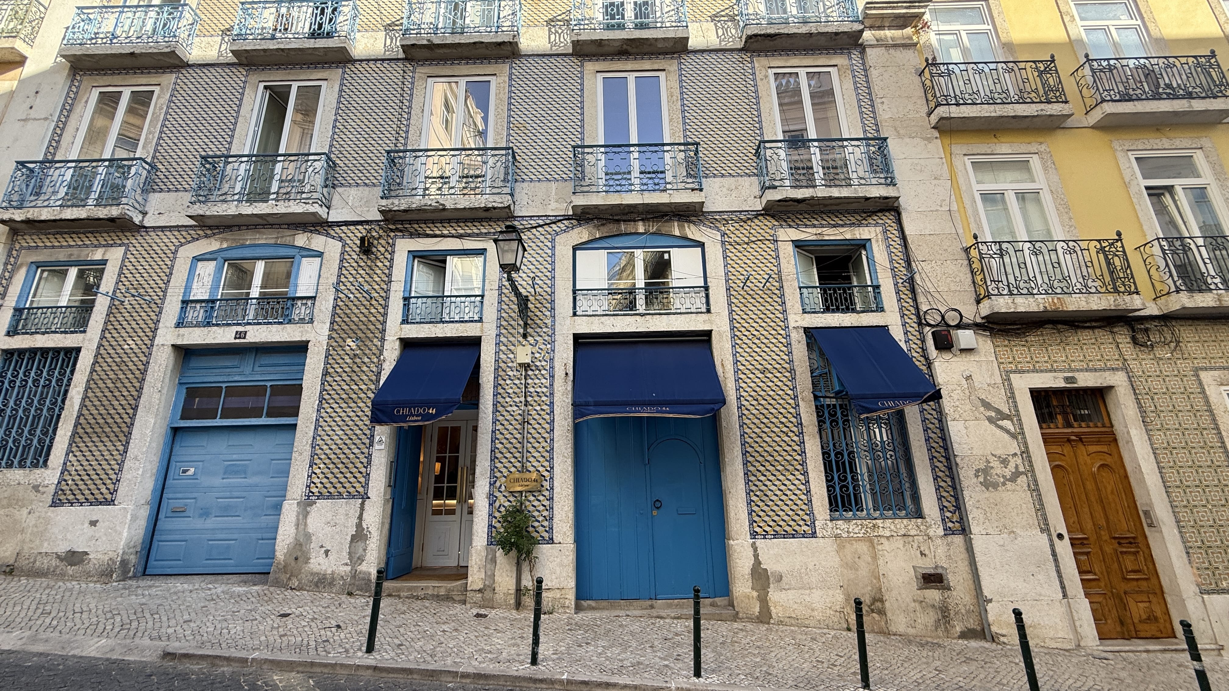 Blue-tiled façade of Chiado 44 Hotel in Lisbon with arched windows, wrought iron balconies, and bold blue doors beneath navy awnings.
