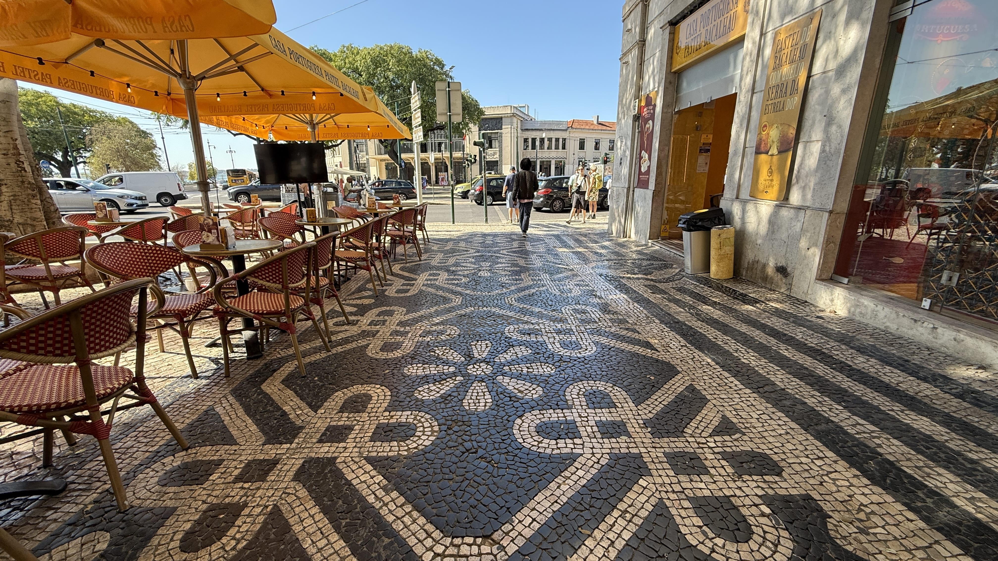 Outdoor café with red chairs and yellow umbrellas on a Lisbon street, featuring traditional black-and-white calçada portuguesa mosaic pavement.