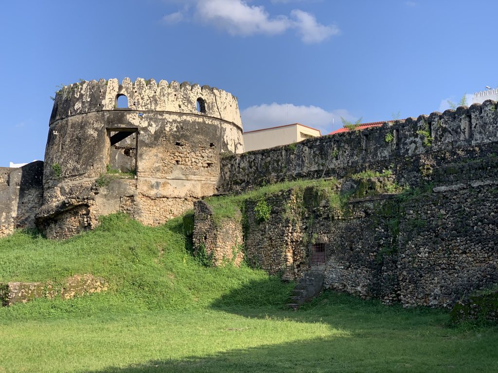 The weathered stone walls and rounded towers of the Old Fort in Stone Town, Zanzibar, surrounded by green grass.