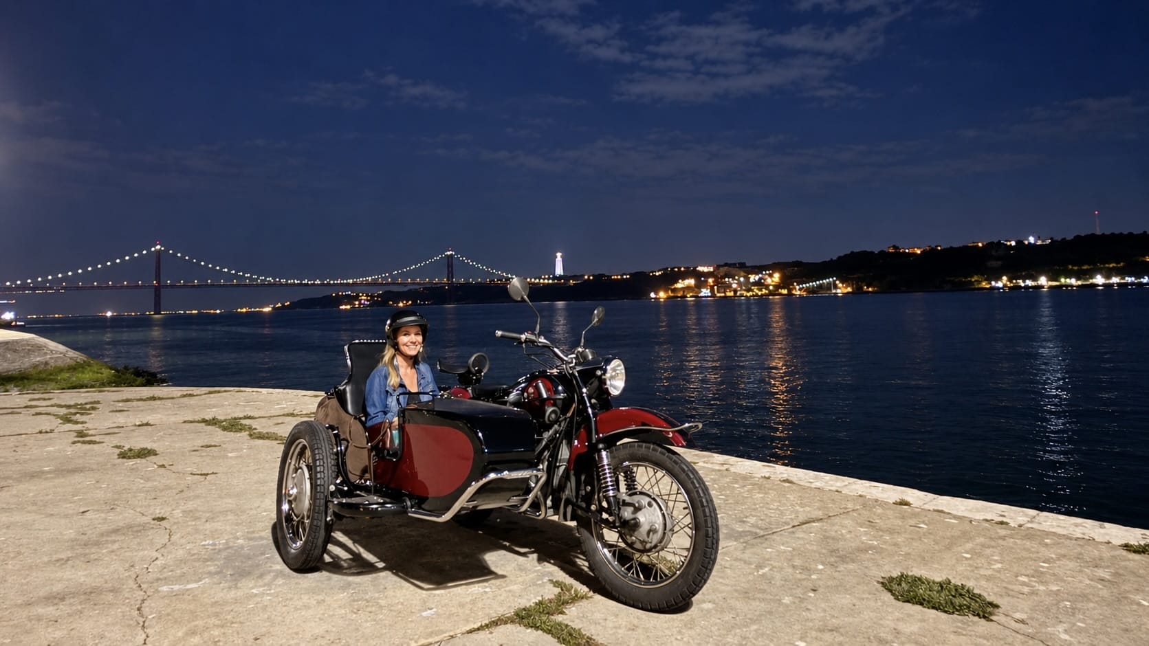 Woman seated in a vintage motorcycle sidecar beside the Tagus River with Lisbon’s illuminated bridge in the background.