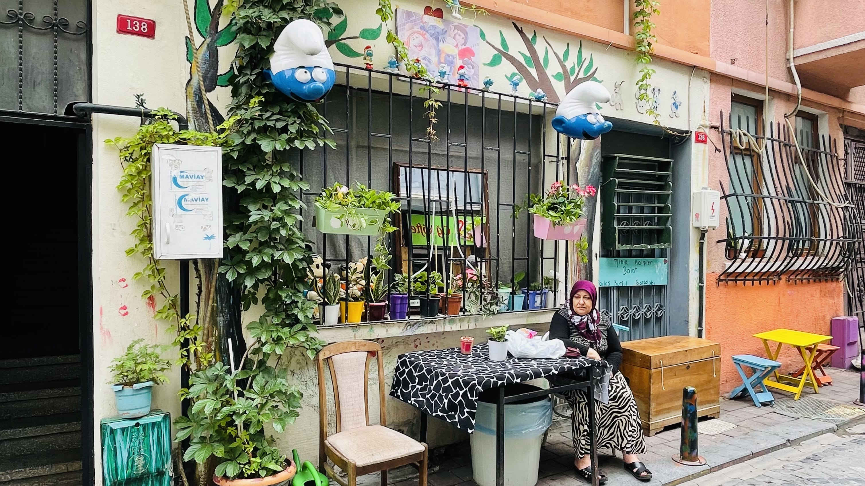 Colorful street scene in Balat, Istanbul, featuring a woman sitting outside a decorated home with potted plants, hanging flower boxes, and playful Smurf decorations above the windows.