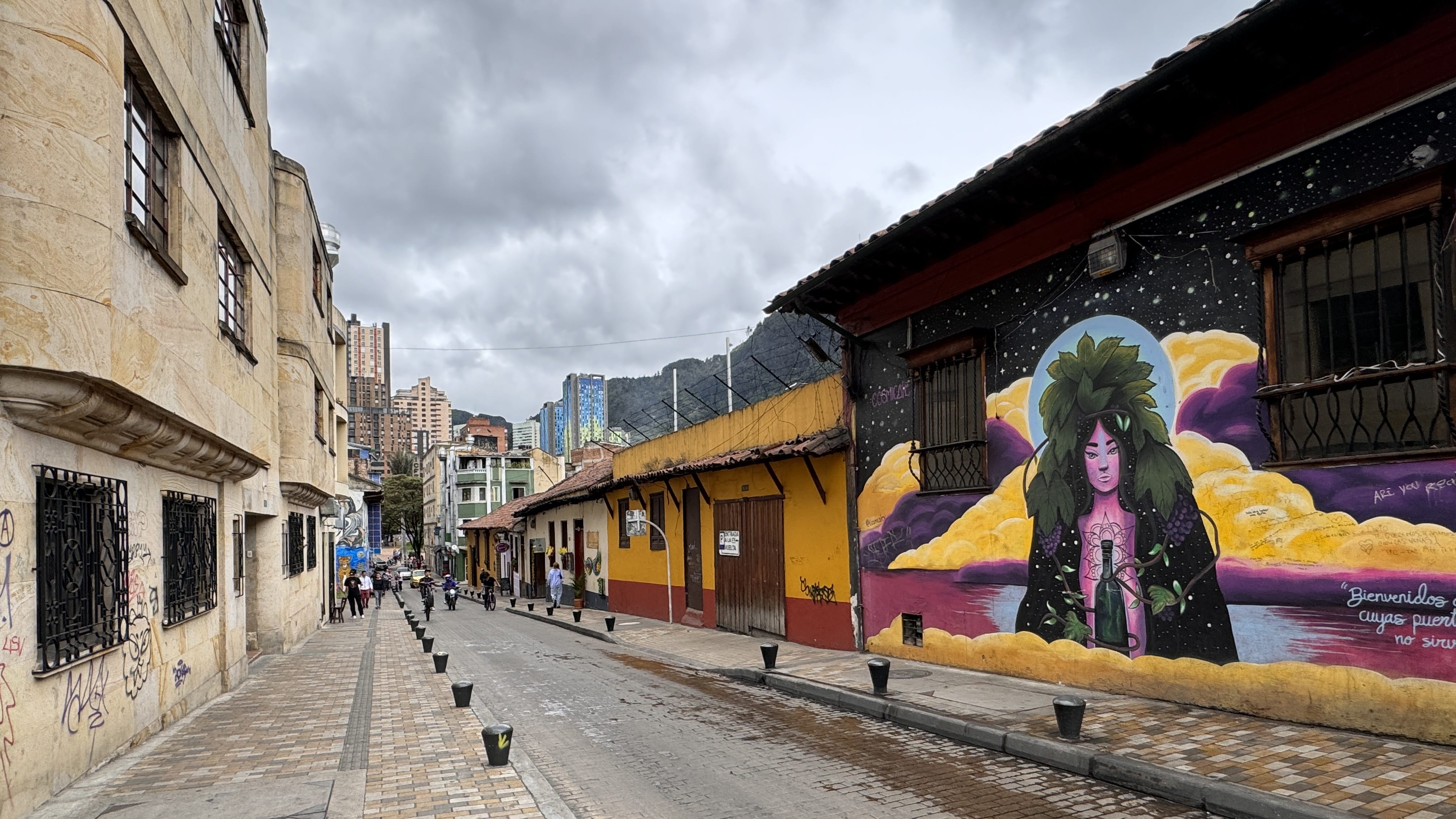 Street art mural of a woman with green leafy hair holding a bottle, painted against a night sky and yellow clouds, in Bogotá’s La Candelaria neighborhood.