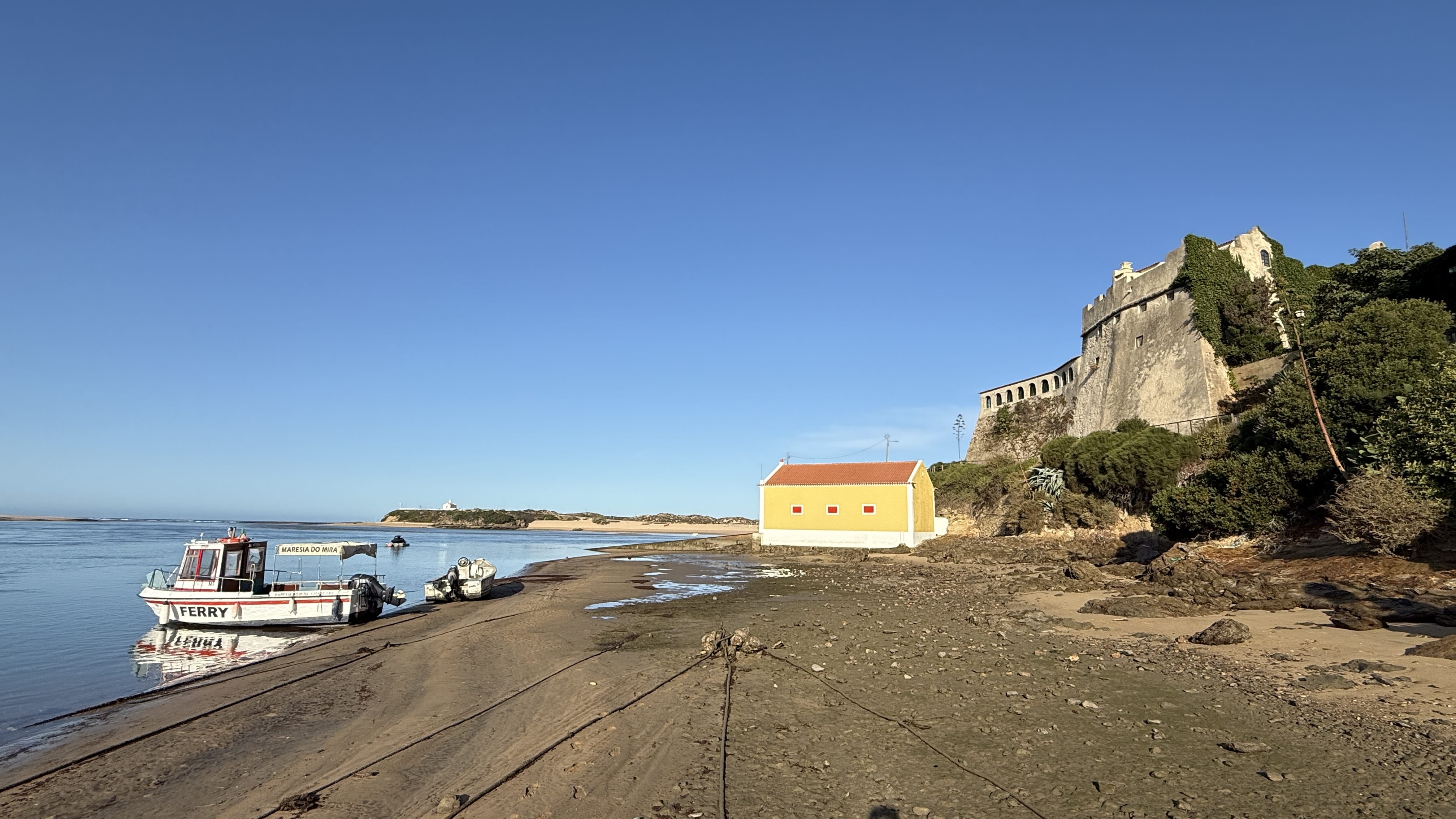 Forte de São Clemente and colorful houses seen from the Mira River in Vila Nova de Milfontes, Portugal, with small boats anchored at the shore.