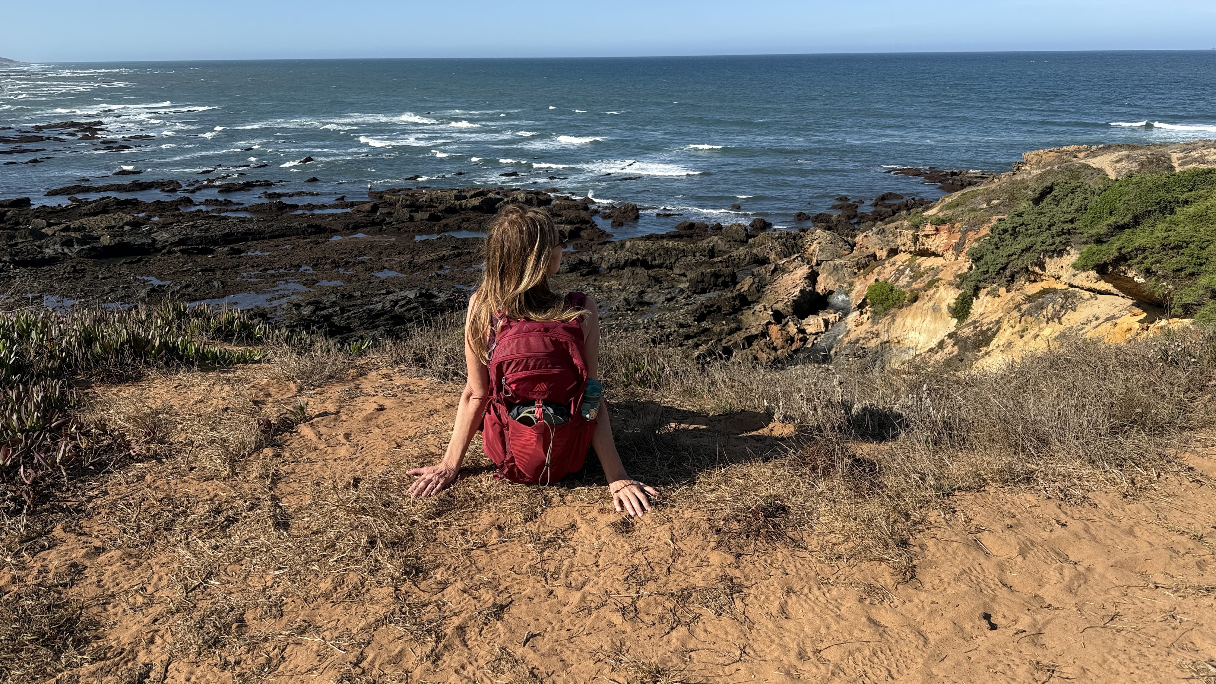 A hiker with a red backpack sitting on a cliff edge along the Fisherman’s Trail in Portugal, looking out at the Atlantic Ocean.
