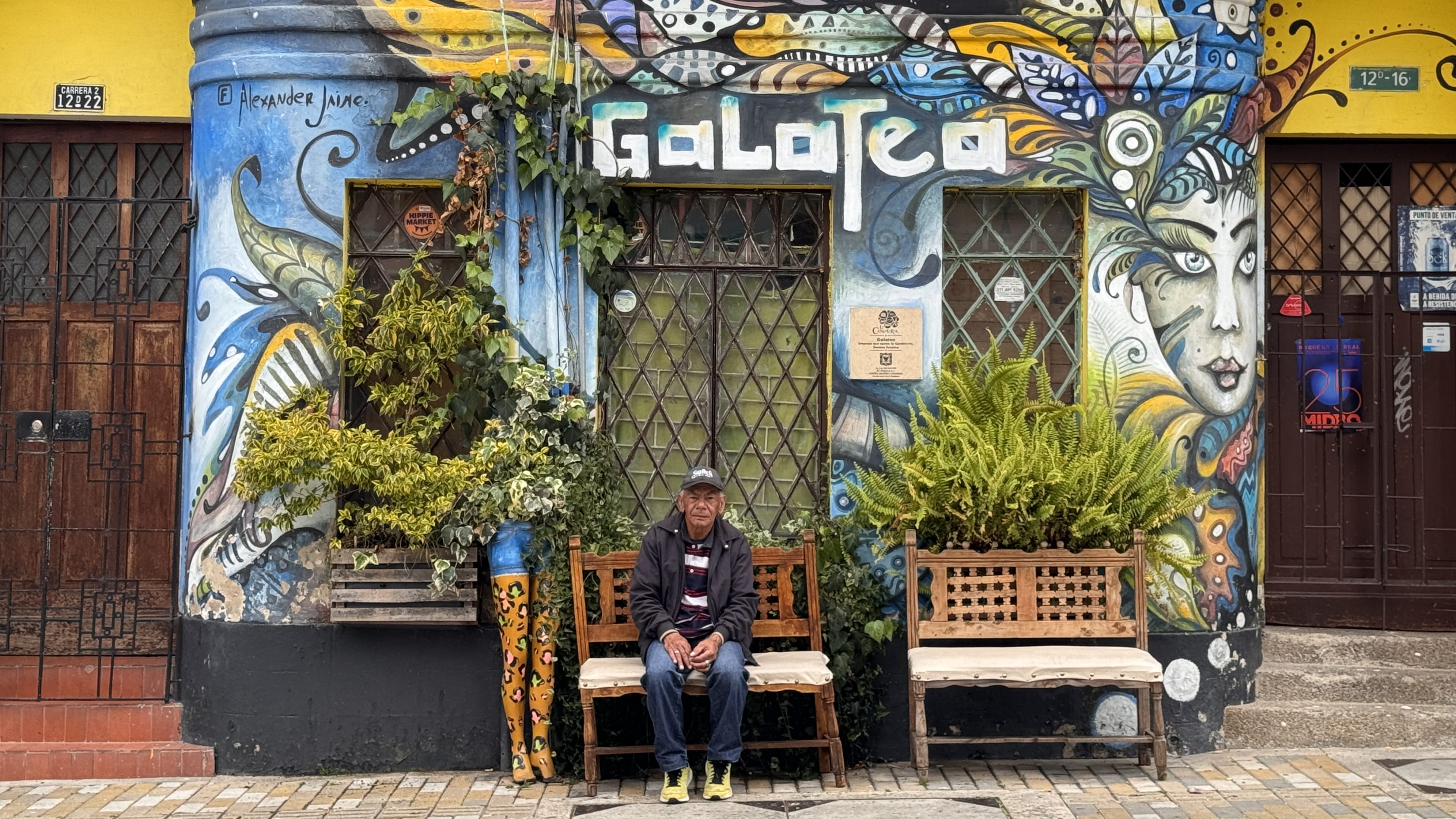 Elderly man sitting on a bench outside a vividly painted building with the word “Galatea” and colorful mural designs in Bogotá, Colombia.
