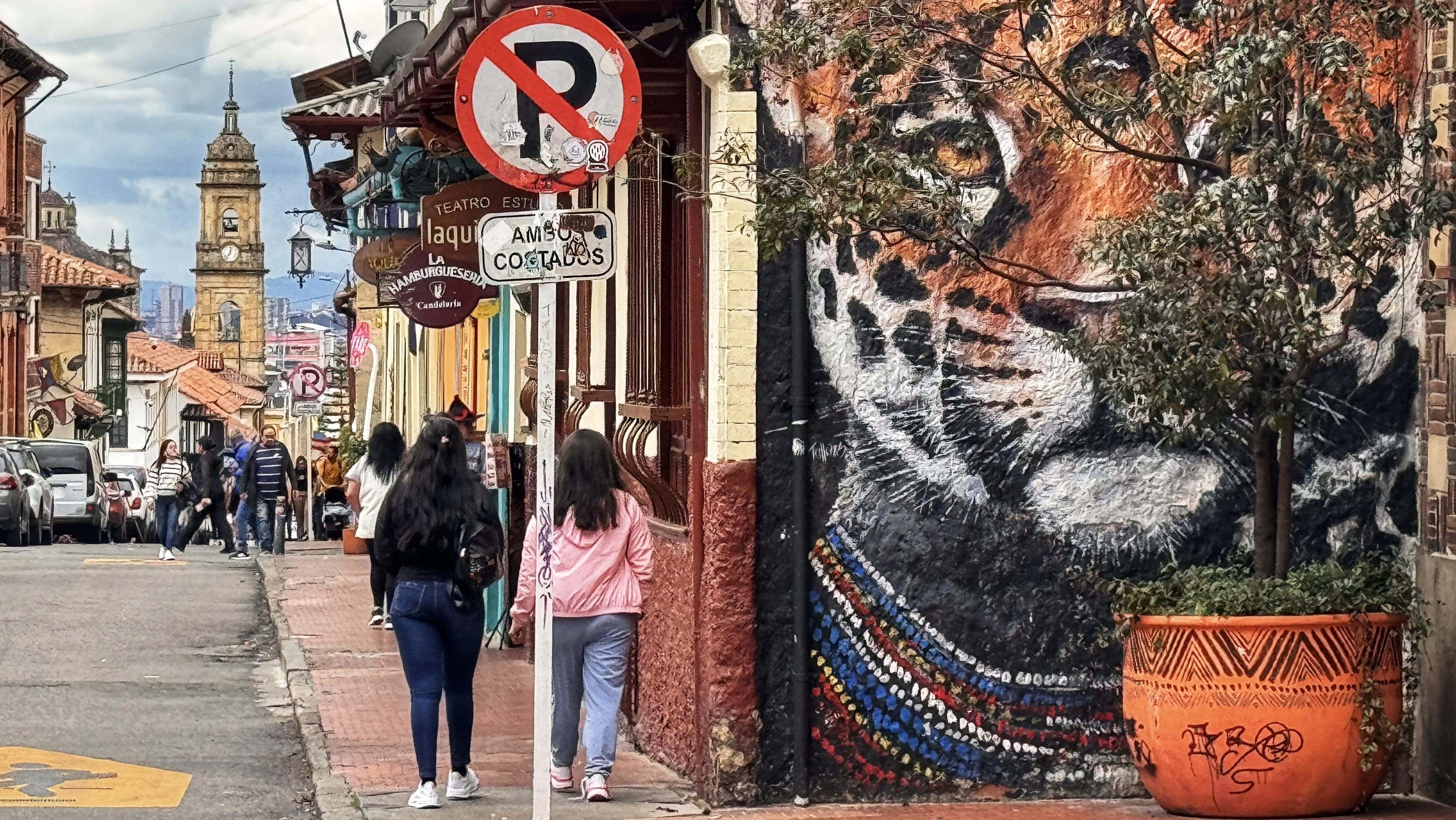 Colorful jaguar mural painted on a building wall in Bogotá’s La Candelaria district, with people walking down the cobblestone street and a colonial clock tower in the distance.