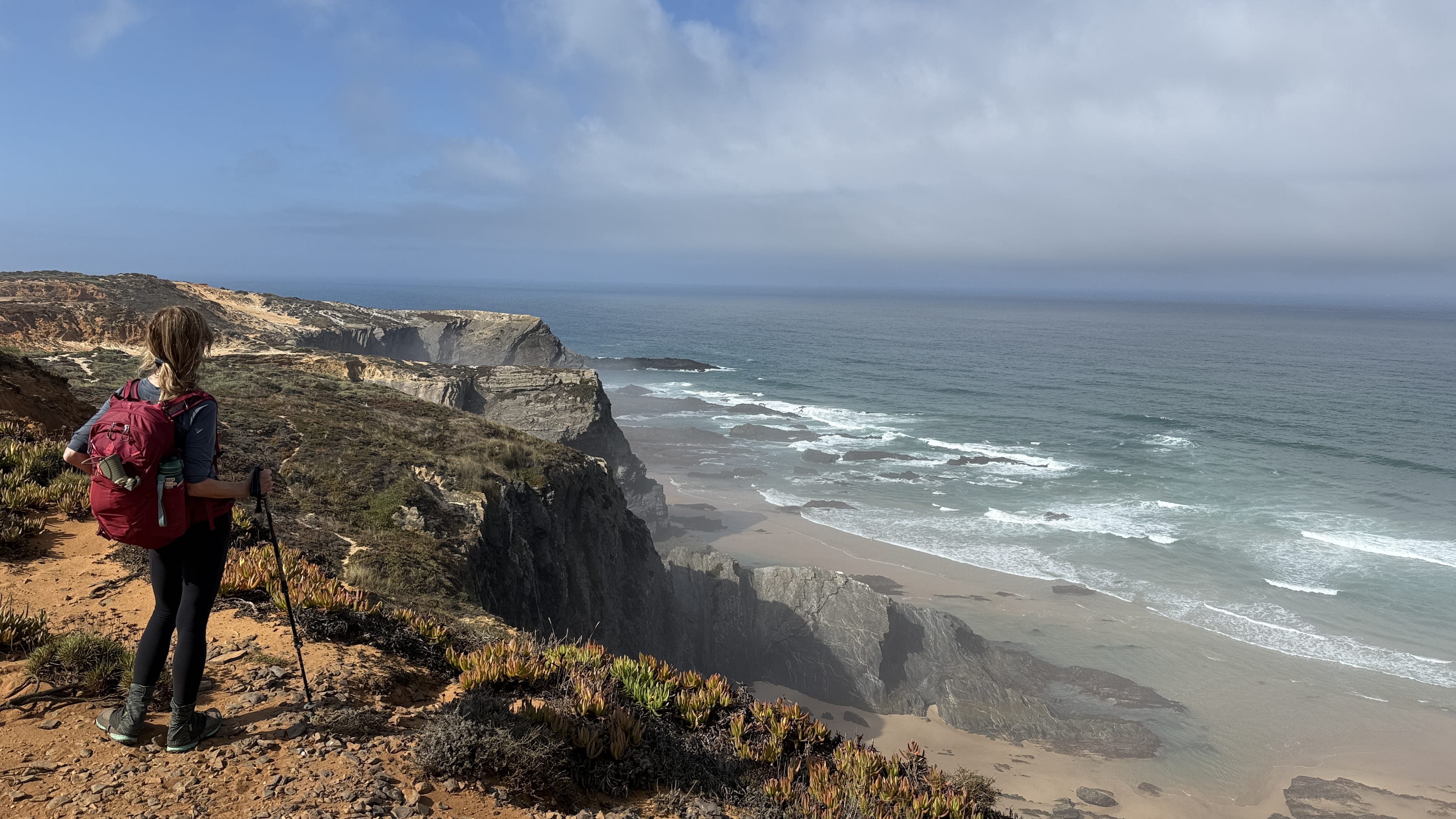Trekker pausing with poles at a clifftop lookout above surf-carved bays on Portugal’s southwest coast.