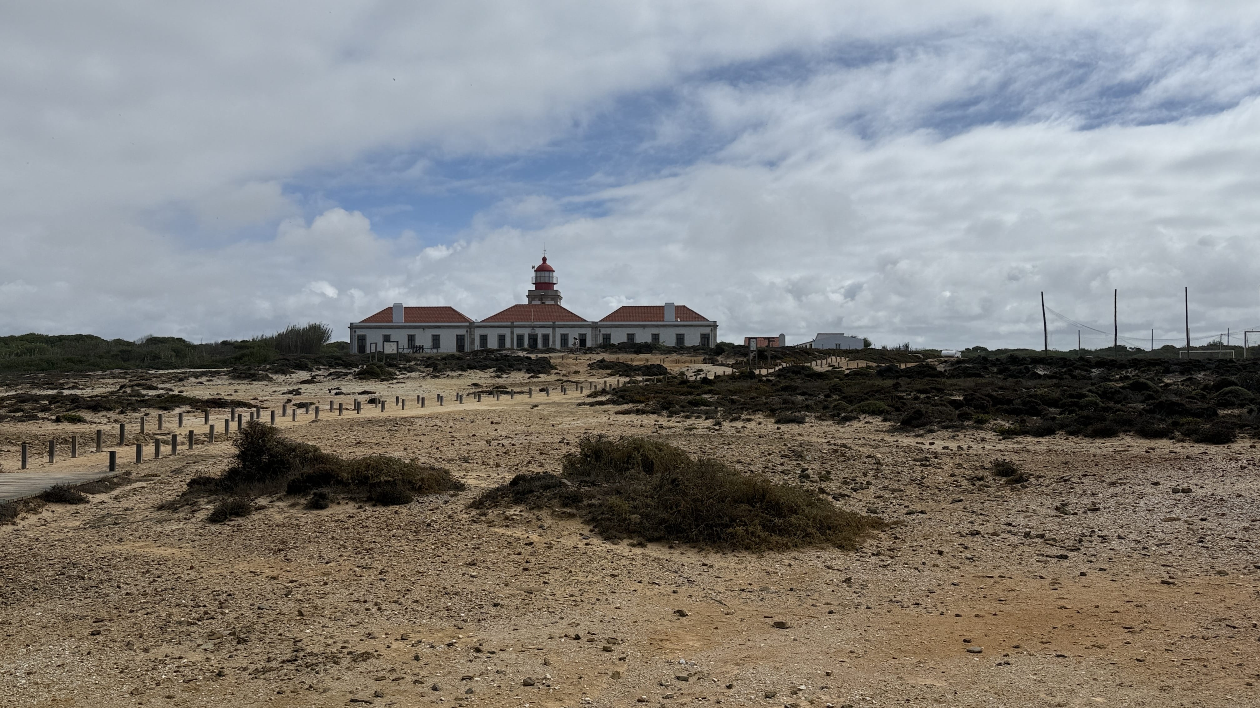 Lighthouse of Cabo Sardão with red dome roof seen from sandy coastal trail under a cloudy sky on the Fishermen’s Trail, Portugal.