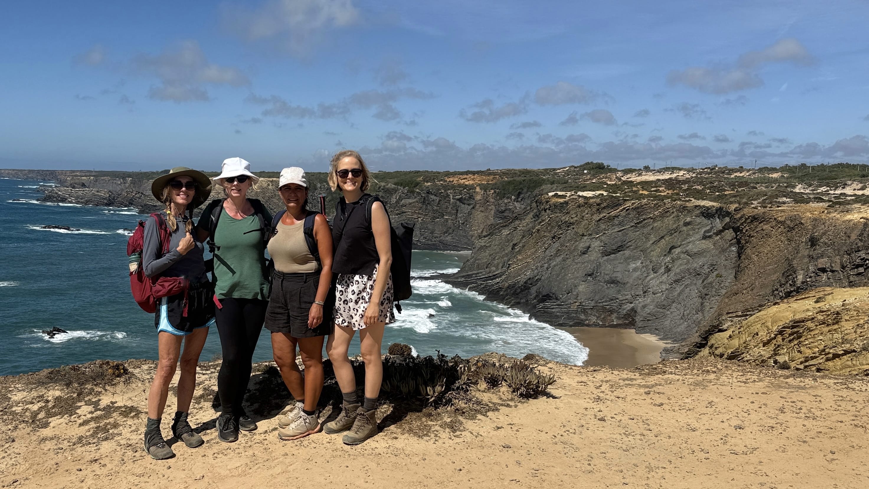 Four women hikers posing on a cliffside viewpoint overlooking the rugged Alentejo coast in Portugal.