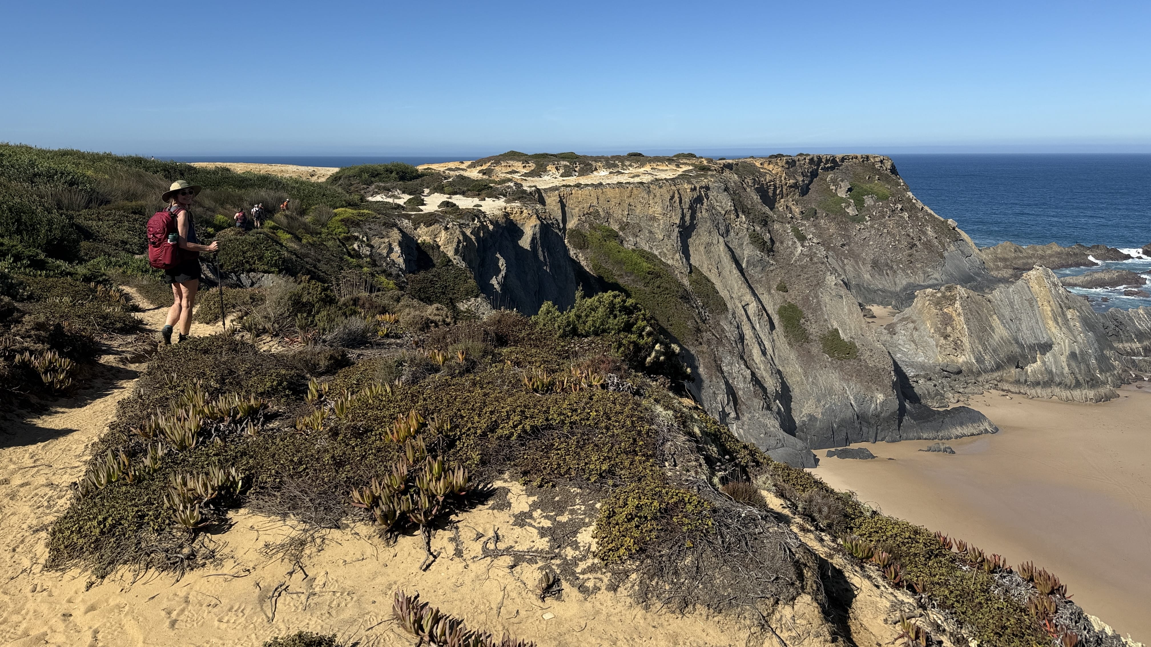 Hiker with red backpack walking along a sandy cliff path above rugged rock formations on Day 5 of the Fisherman’s Trail, Portugal.