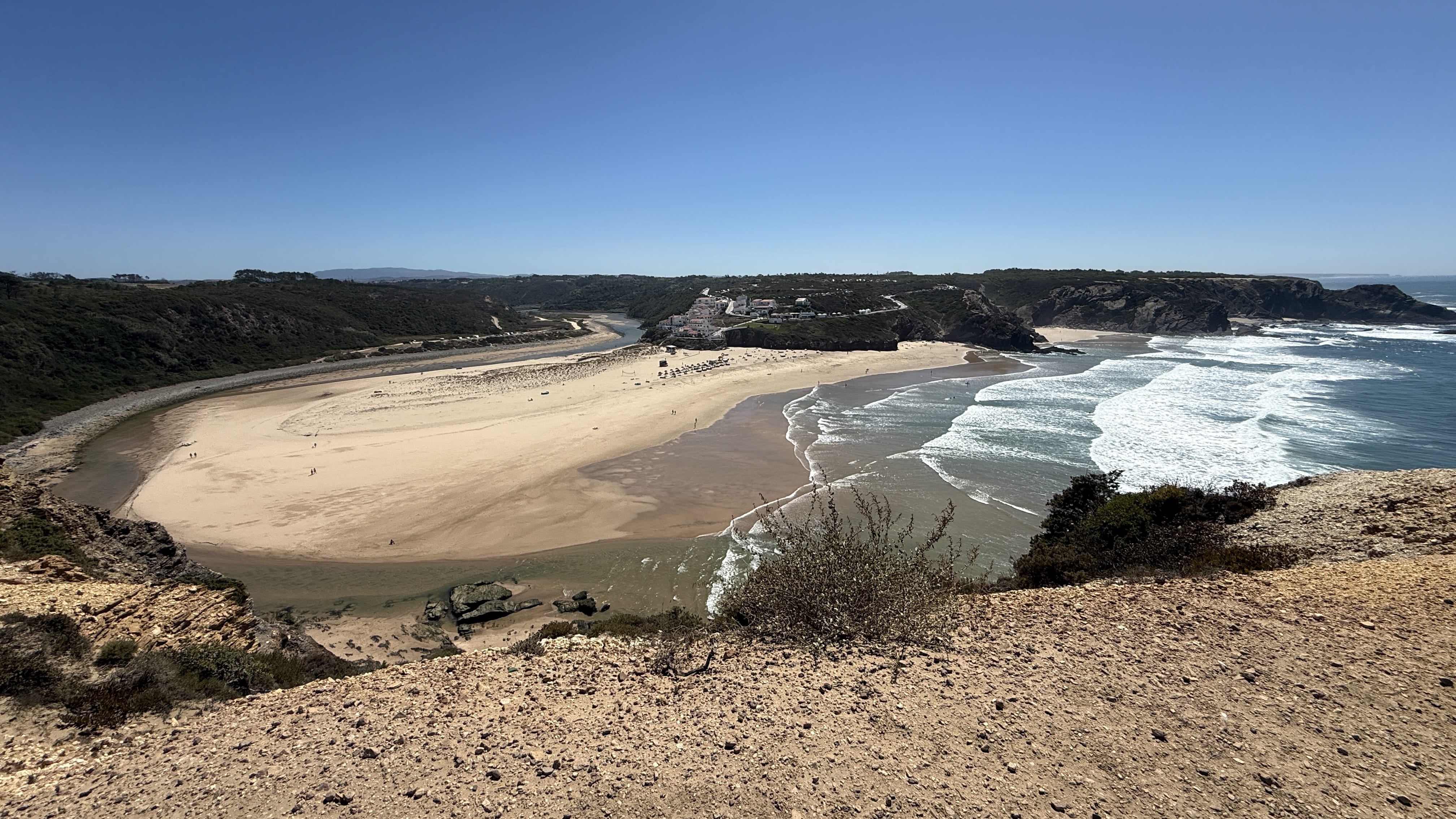 Aerial-style view of Praia da Baiona, with golden sand stretching between the river mouth and the Atlantic Ocean, framed by cliffs and the whitewashed village of Odeceixe in the distance.