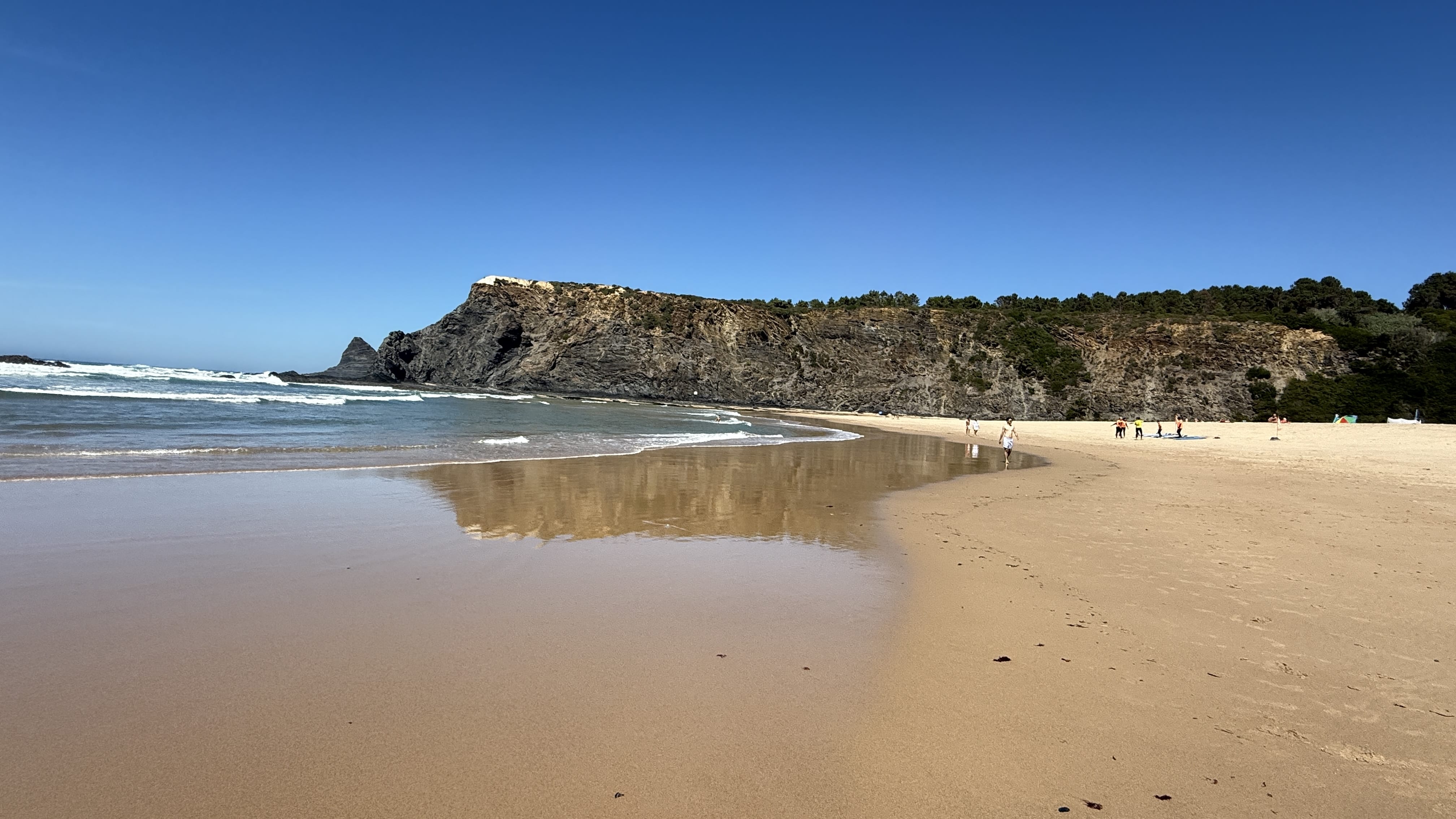 Sandy stretch of Praia da Baiona with gentle waves lapping the shore, cliffs rising in the background, and people walking along the beach.