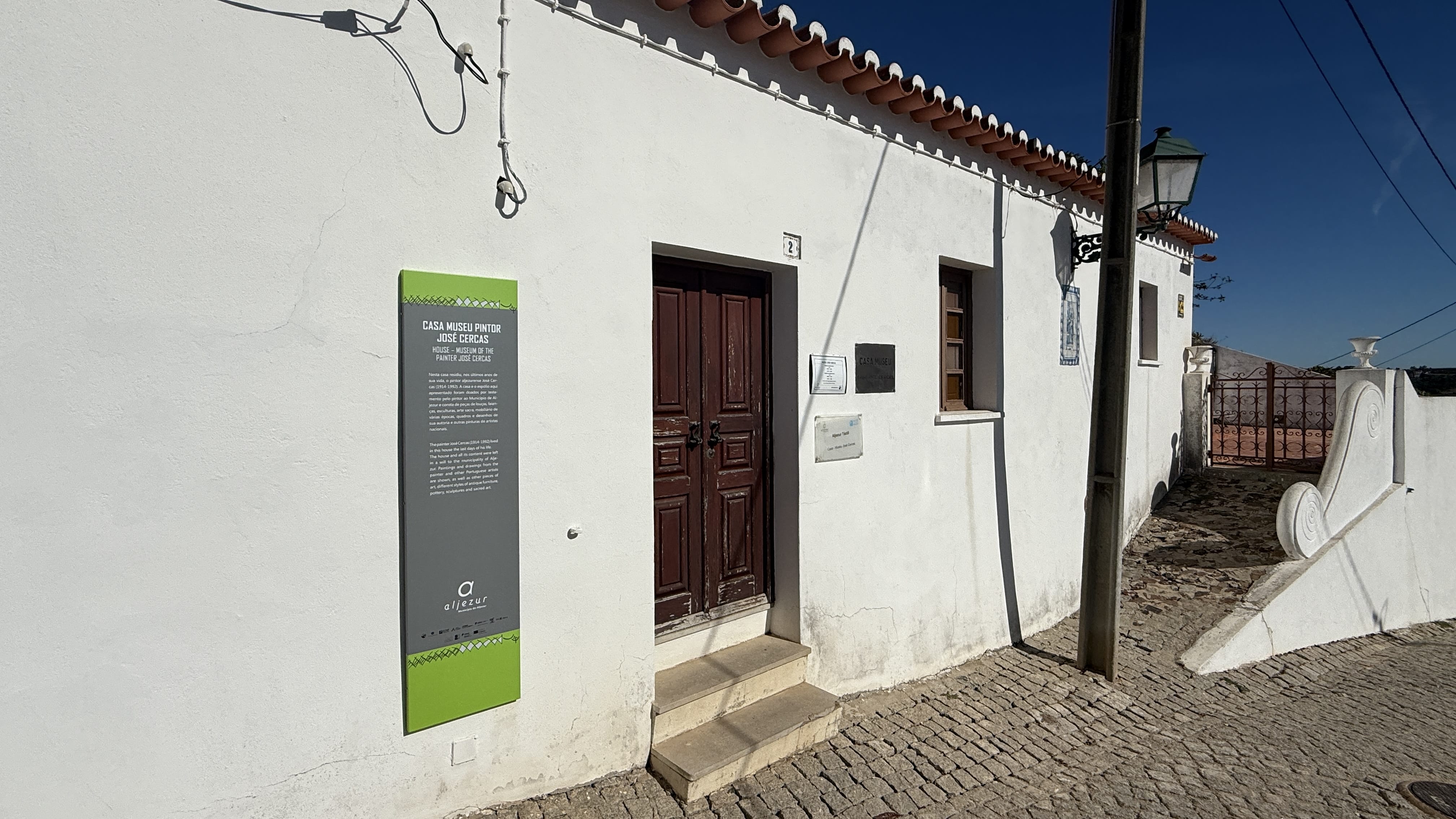 : Whitewashed exterior of Casa Museu Pintor José Cercas in Aljezur, Portugal, with a wooden door and museum sign on the wall.