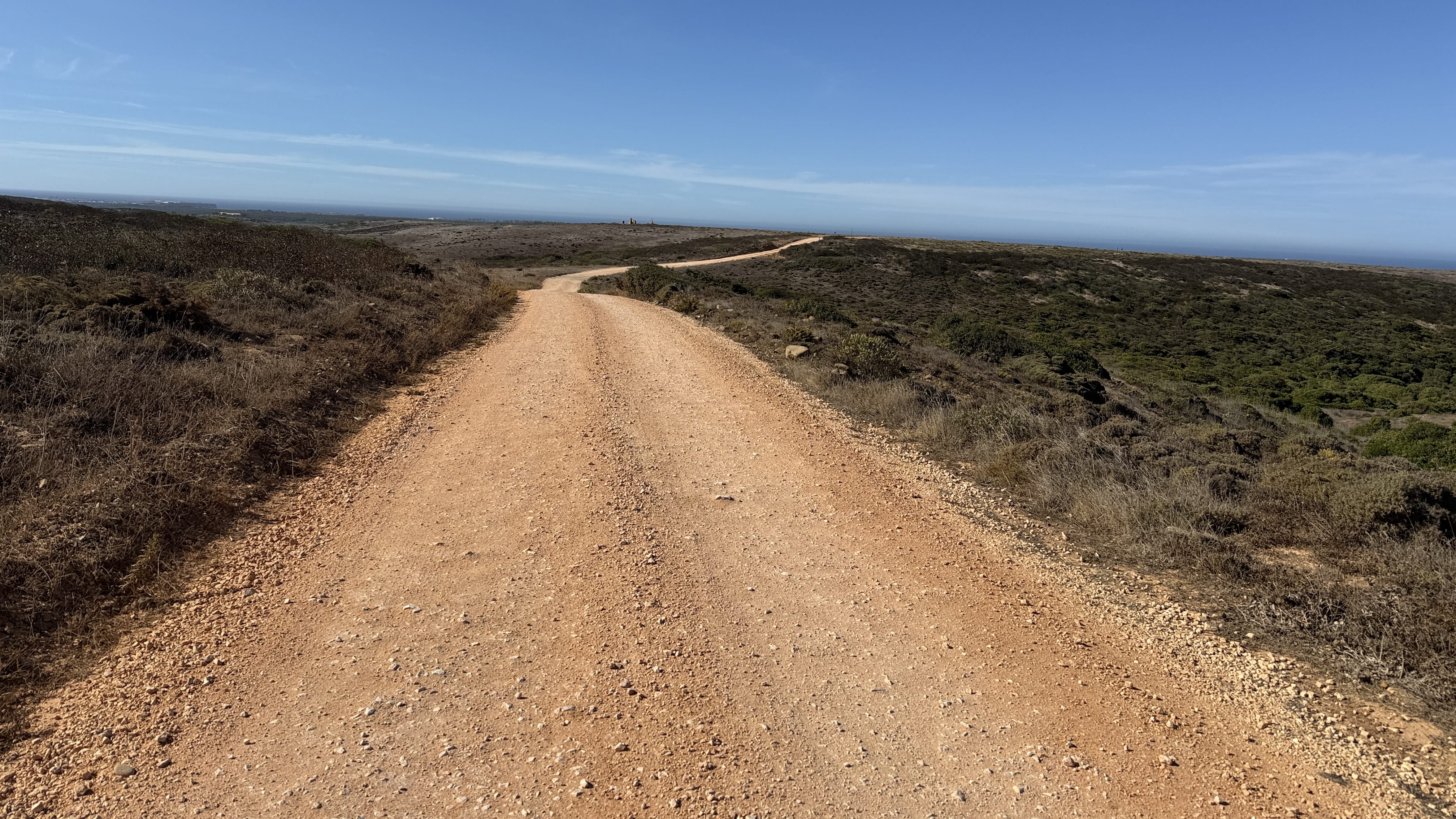 Wide dirt track winding across the dry landscape of the Algarve with ocean visible in the distance.