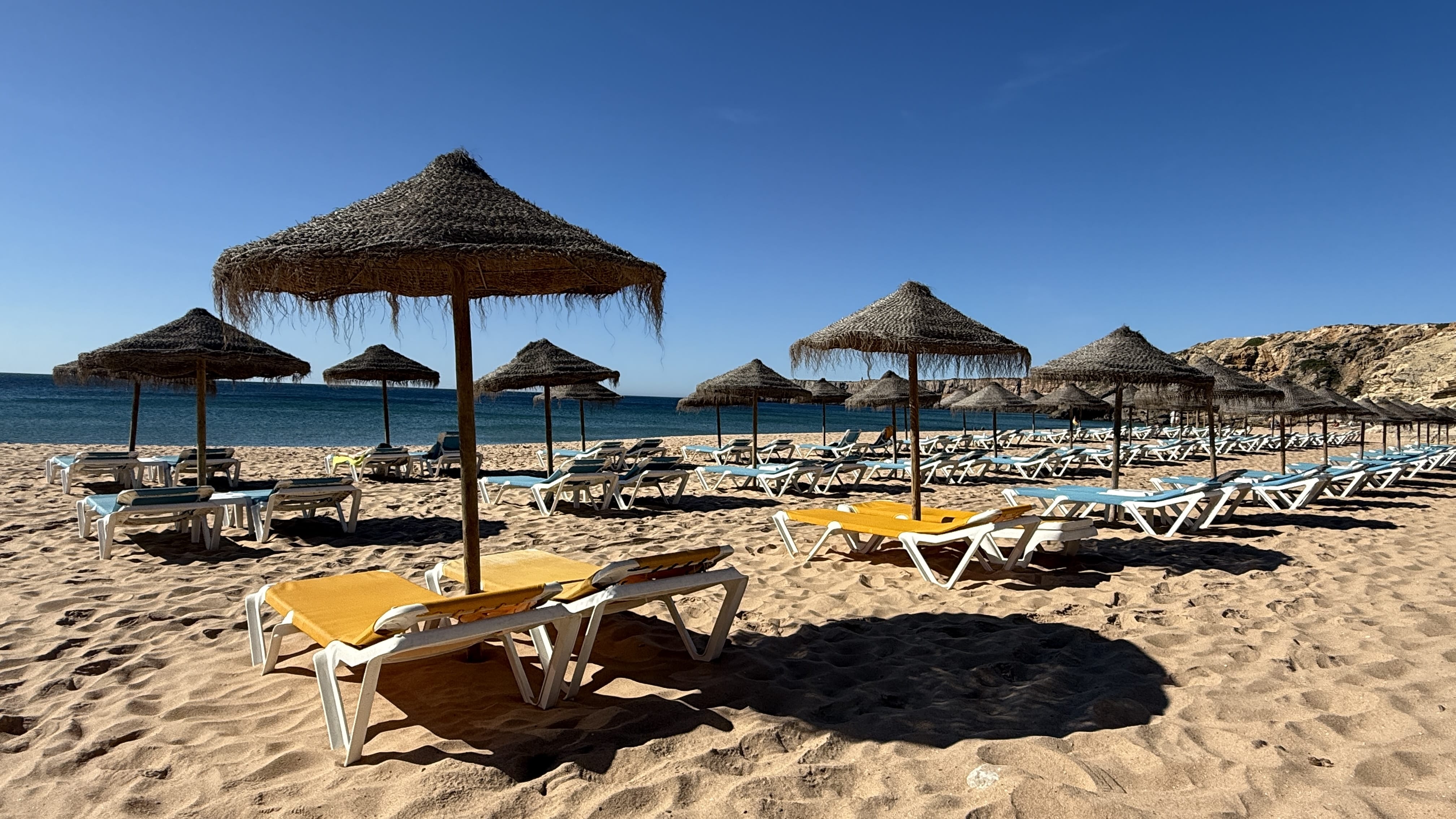 Rows of straw parasols and empty lounge chairs on a sandy beach in Sagres, Portugal, with the blue Atlantic Ocean in the background.