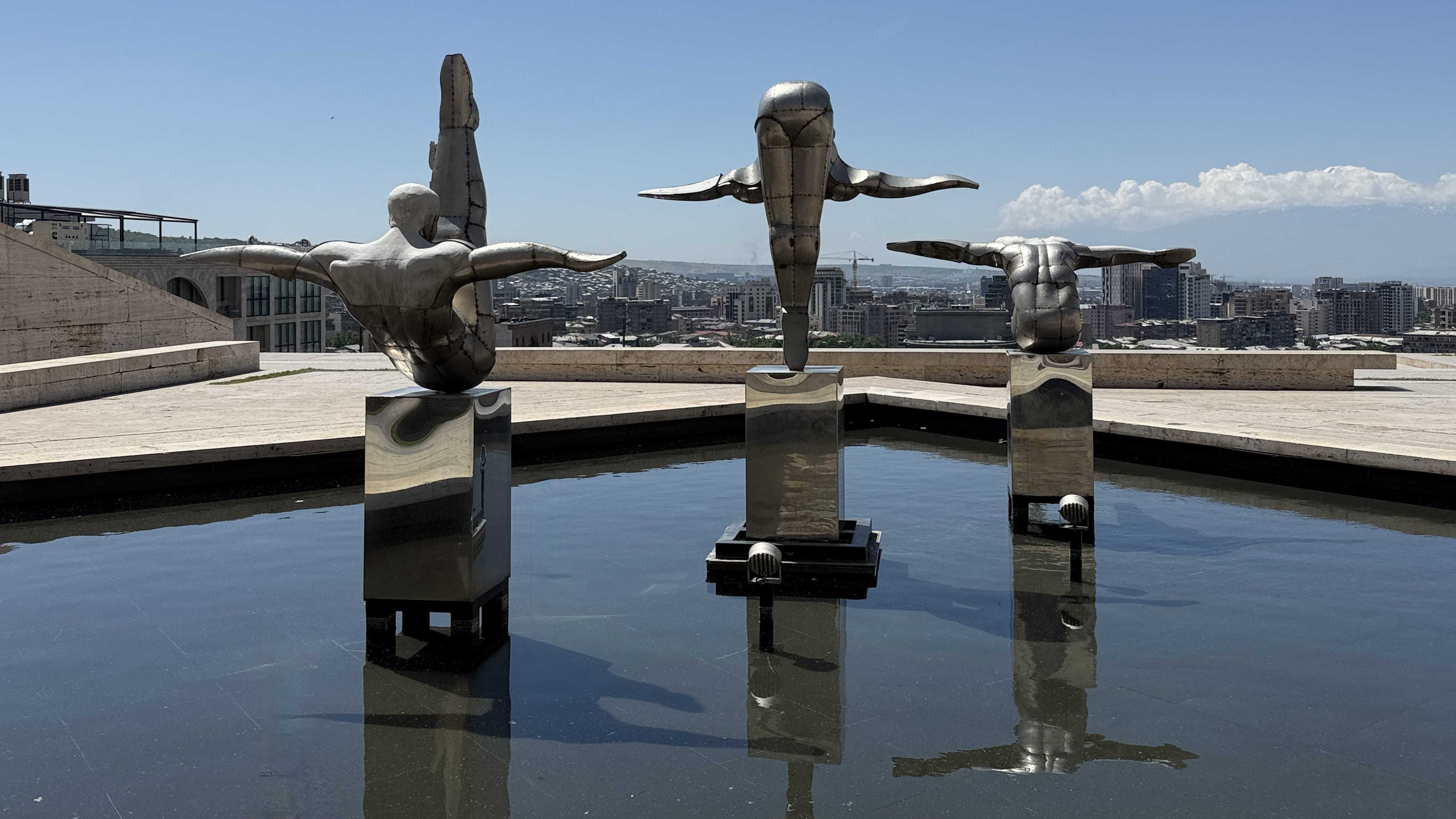 Modern outdoor sculpture of three stainless steel diver figures at the Cascade Complex in Yerevan, Armenia, reflecting in the water with the city and Mount Ararat in the background – 24 hours in Yerevan