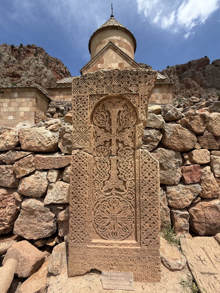 Ornately carved khachkar (Armenian cross-stone) in front of Noravank Monastery in Armenia, with stone church and cliffs in the background.