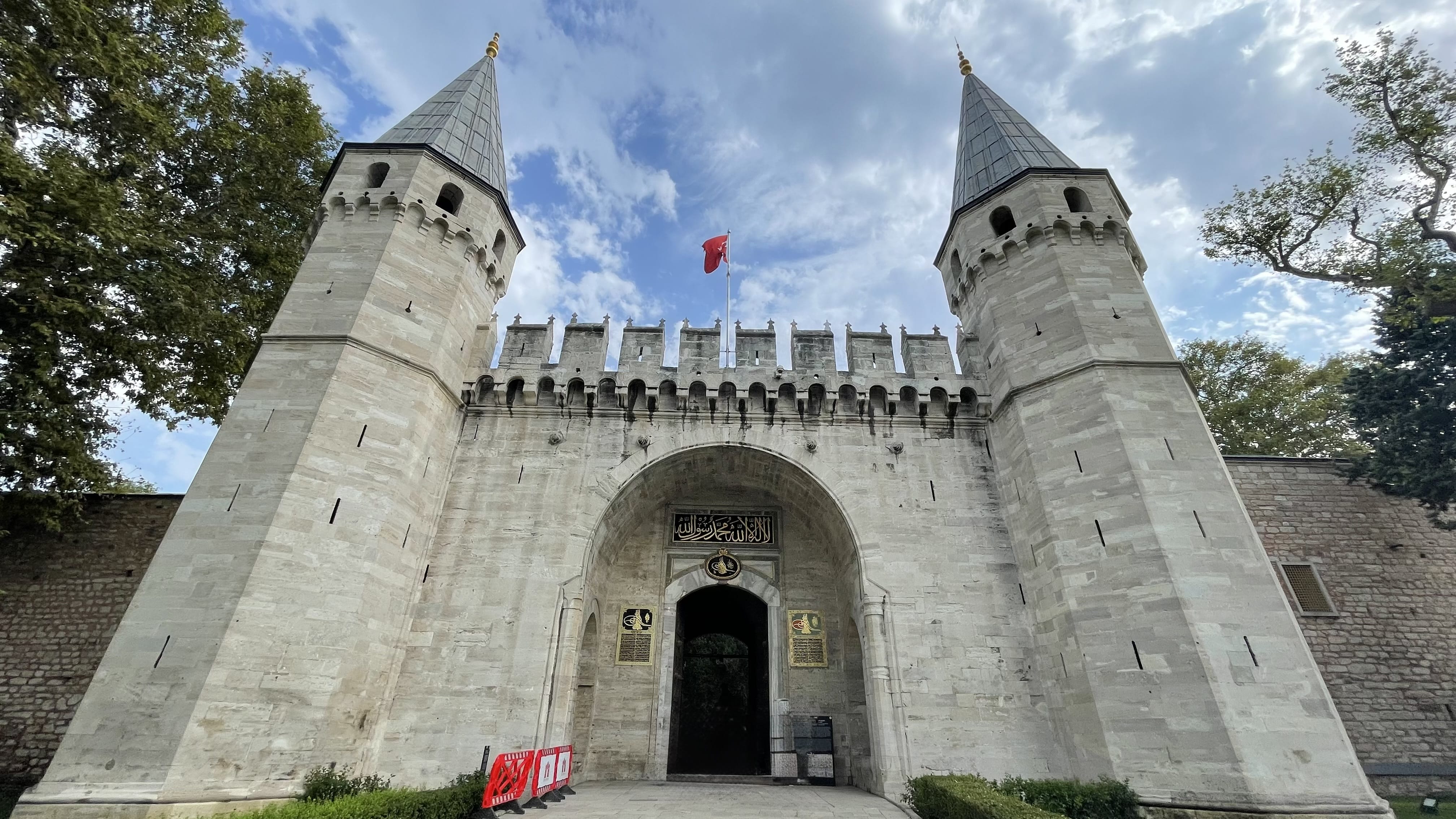 Entrance gate to Topkapi Palace in Istanbul with two tall towers and a Turkish flag flying above.