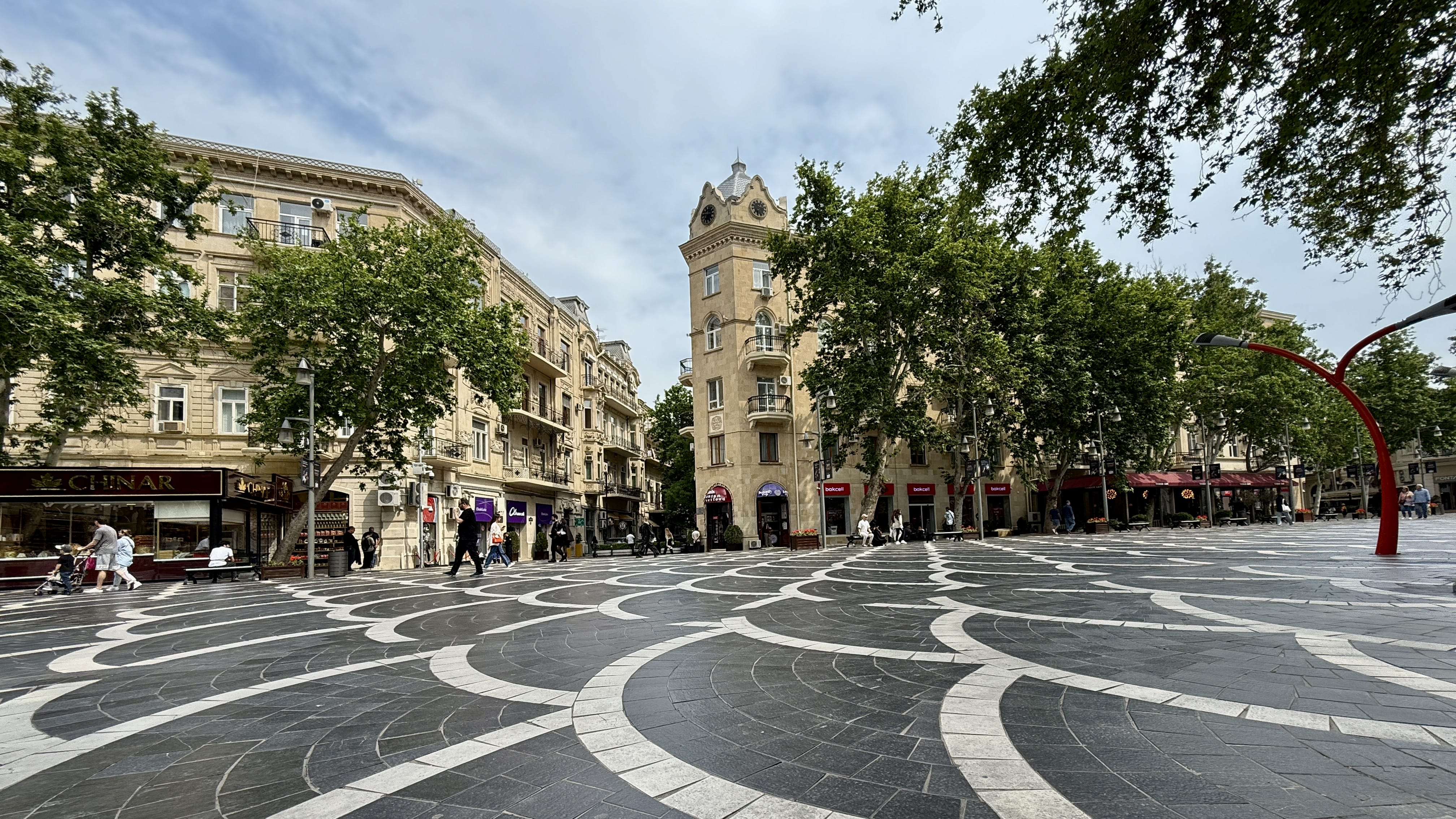 Fountain Square (Fəvvarələr Meydanı) in central Baku, Azerbaijan, featuring black-and-white patterned paving stones, historic sandstone buildings with ornate balconies, trees lining the pedestrian plaza, and people walking near cafés and shops.