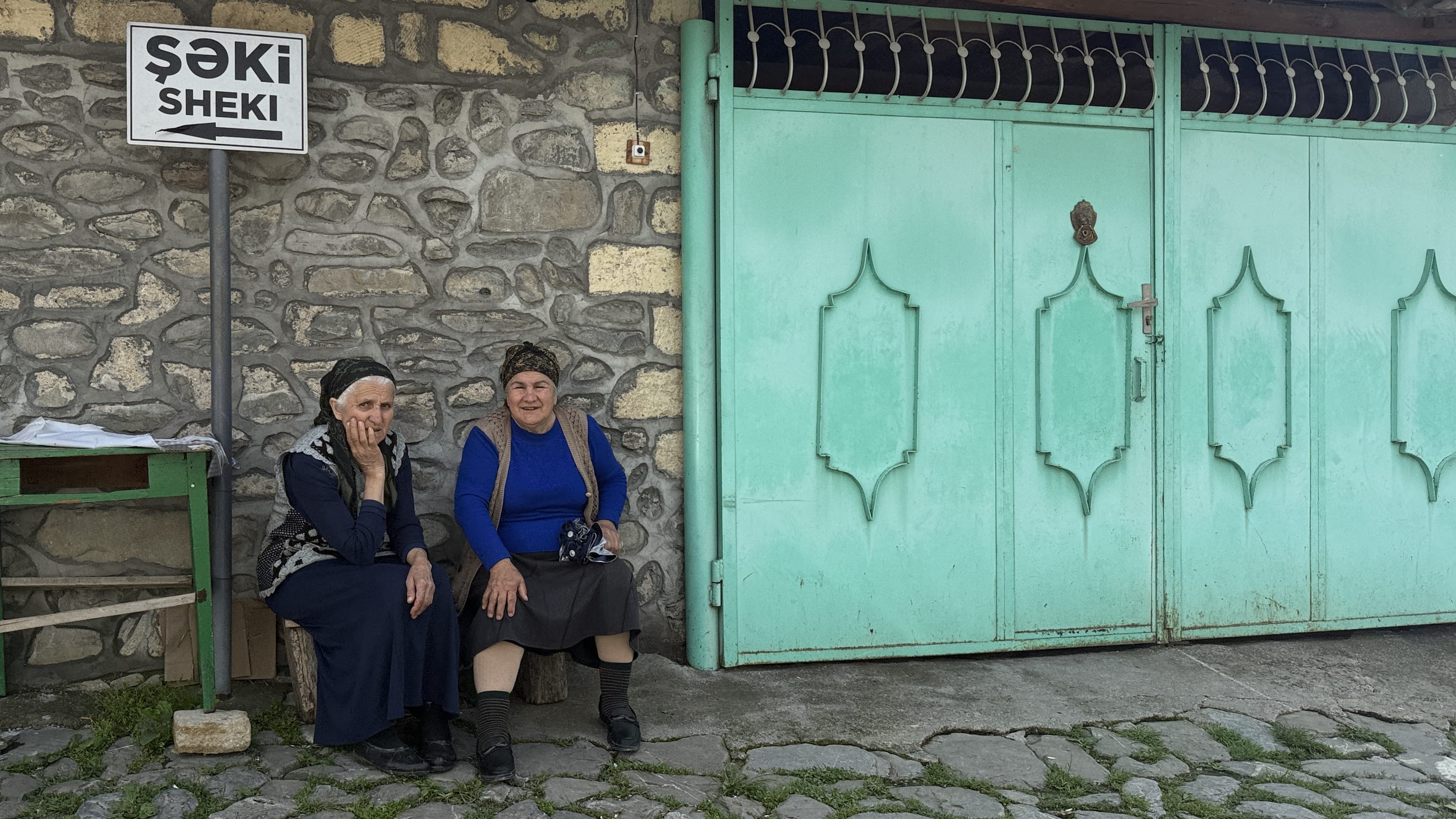 Two local women sitting on a stone street under a Sheki sign, photographed during my 7 Days in Azerbaijan.
