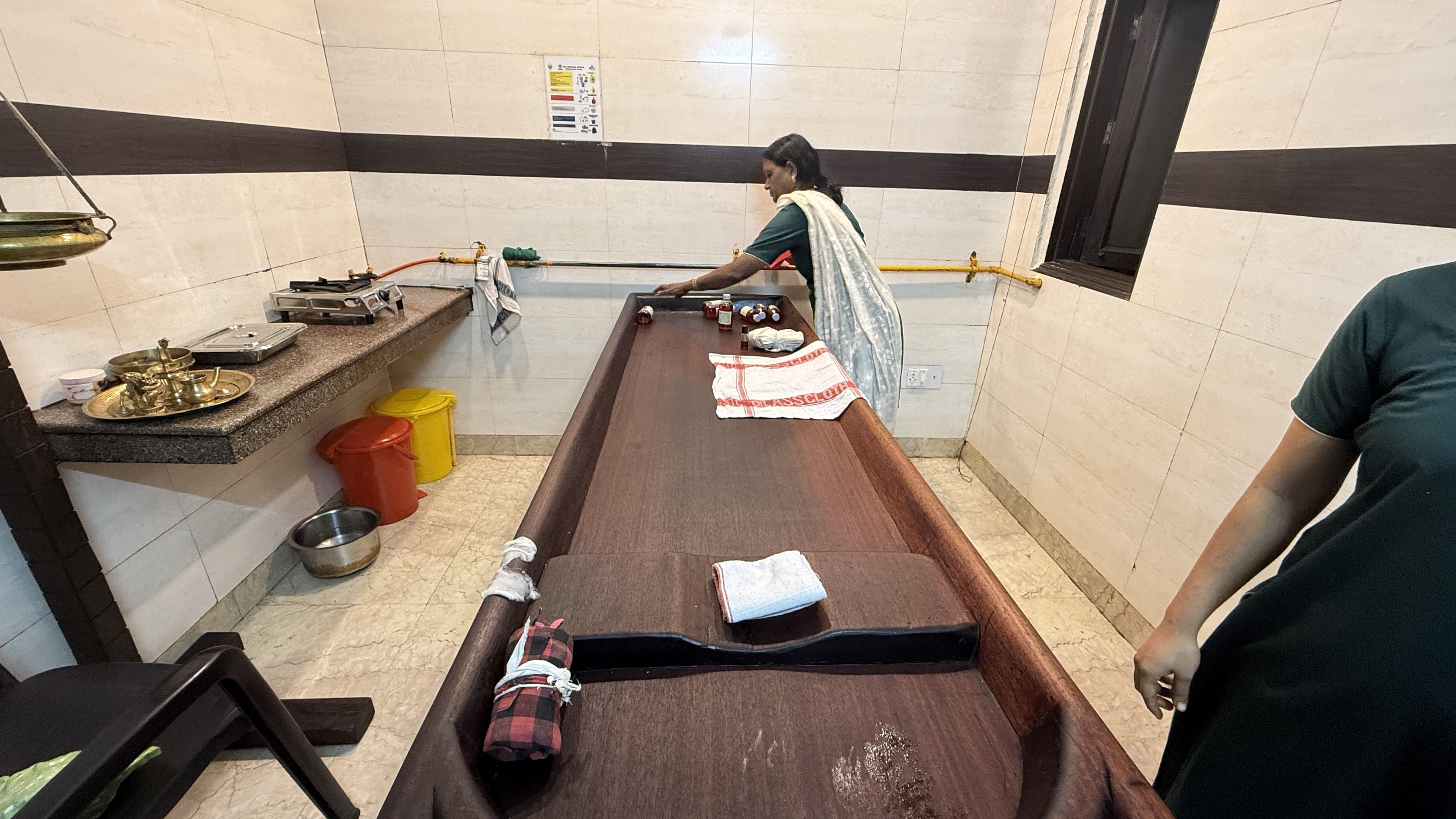 Ayurvedic therapist preparing a traditional wooden droni massage table at Dr. Sudha’s Ayurveda Kendra in Delhi, with oils, boluses, and towels set out for an Abhyangam treatment fro my The outside of Dr. Sudha’s Ayurveda Kendra in South Delhi — not a hotel spa, but a working Ayurvedic clinic where many people come for an Ayurvedic Massage in Delhi —Ayurvedic spa included in this 2 Days in Delhi itinerary