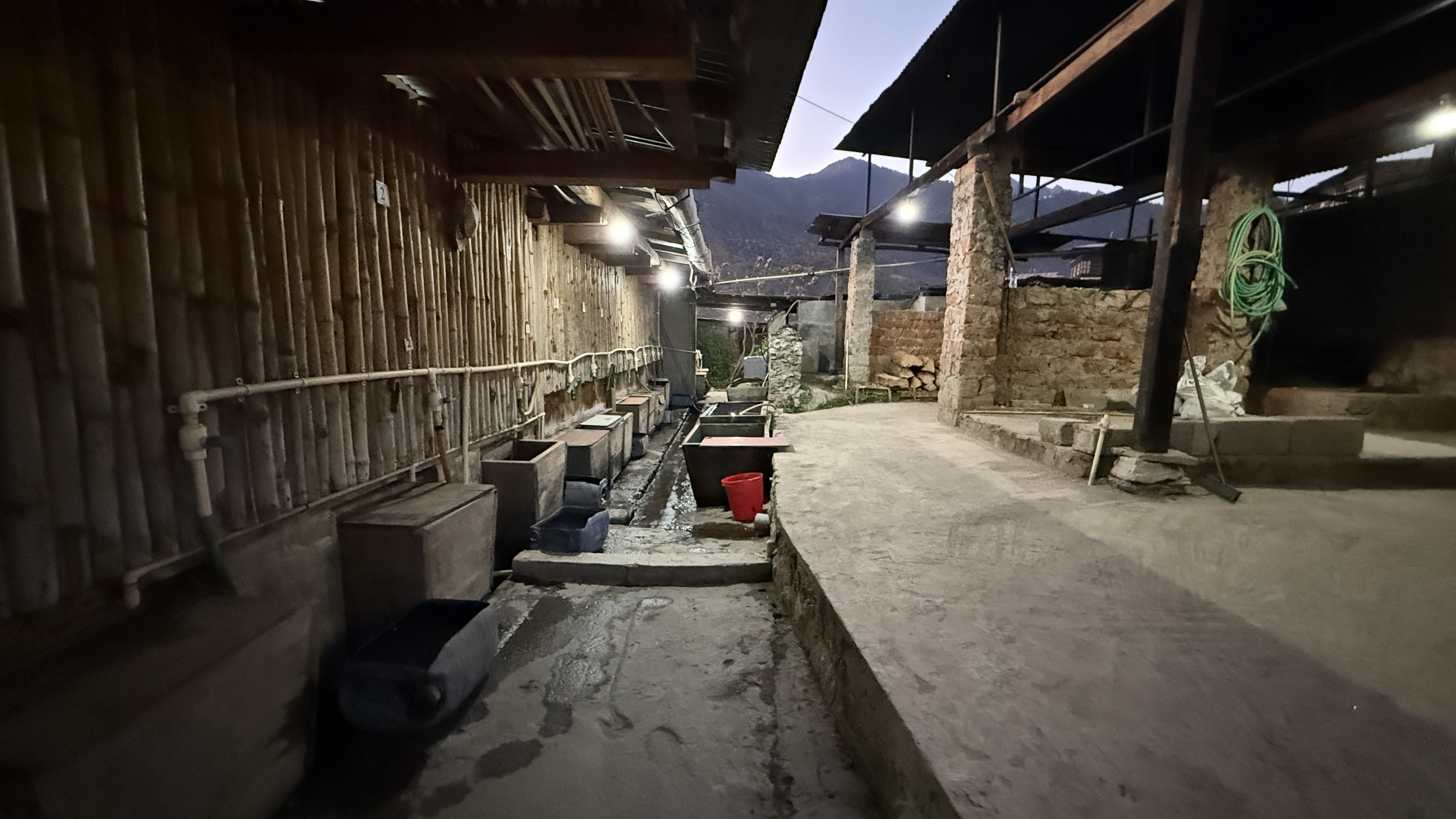Outdoor corridor of a Bhutanese hot stone bath facility with stone tubs, pipes, and metal roofing, set against the mountains at dusk.