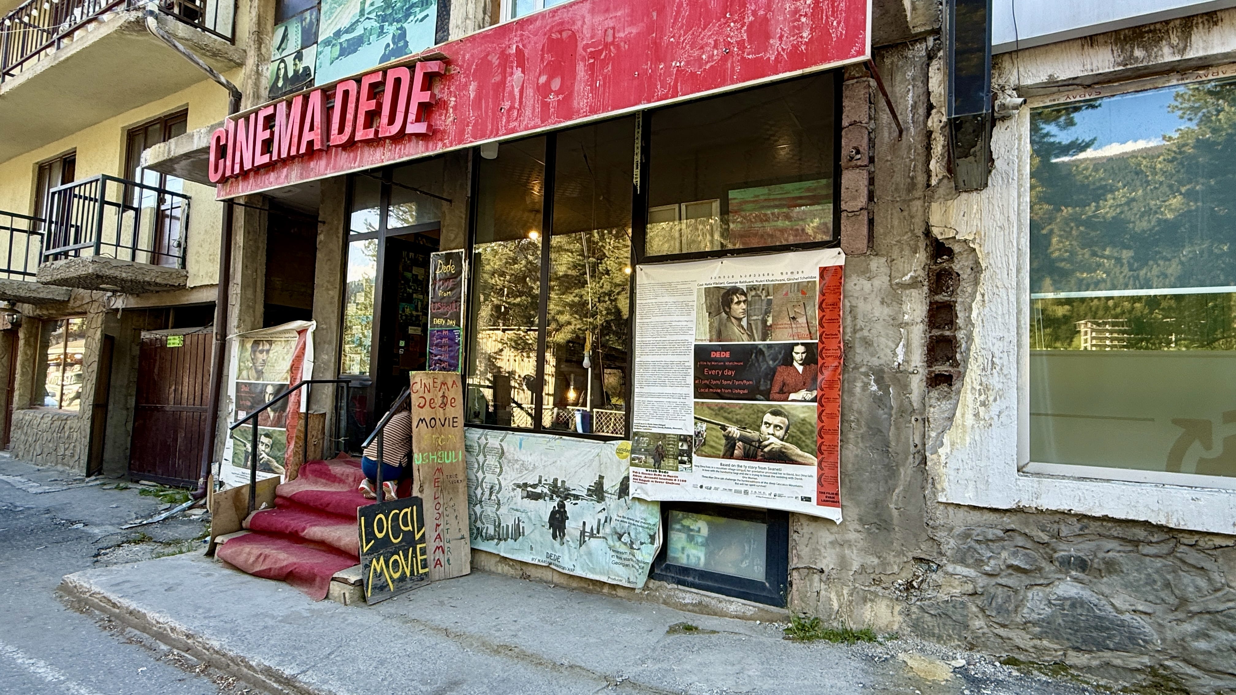 Street view of Dede Cinema entrance in Mestia, Svaneti, Georgia, with posters and a red sign above the door.