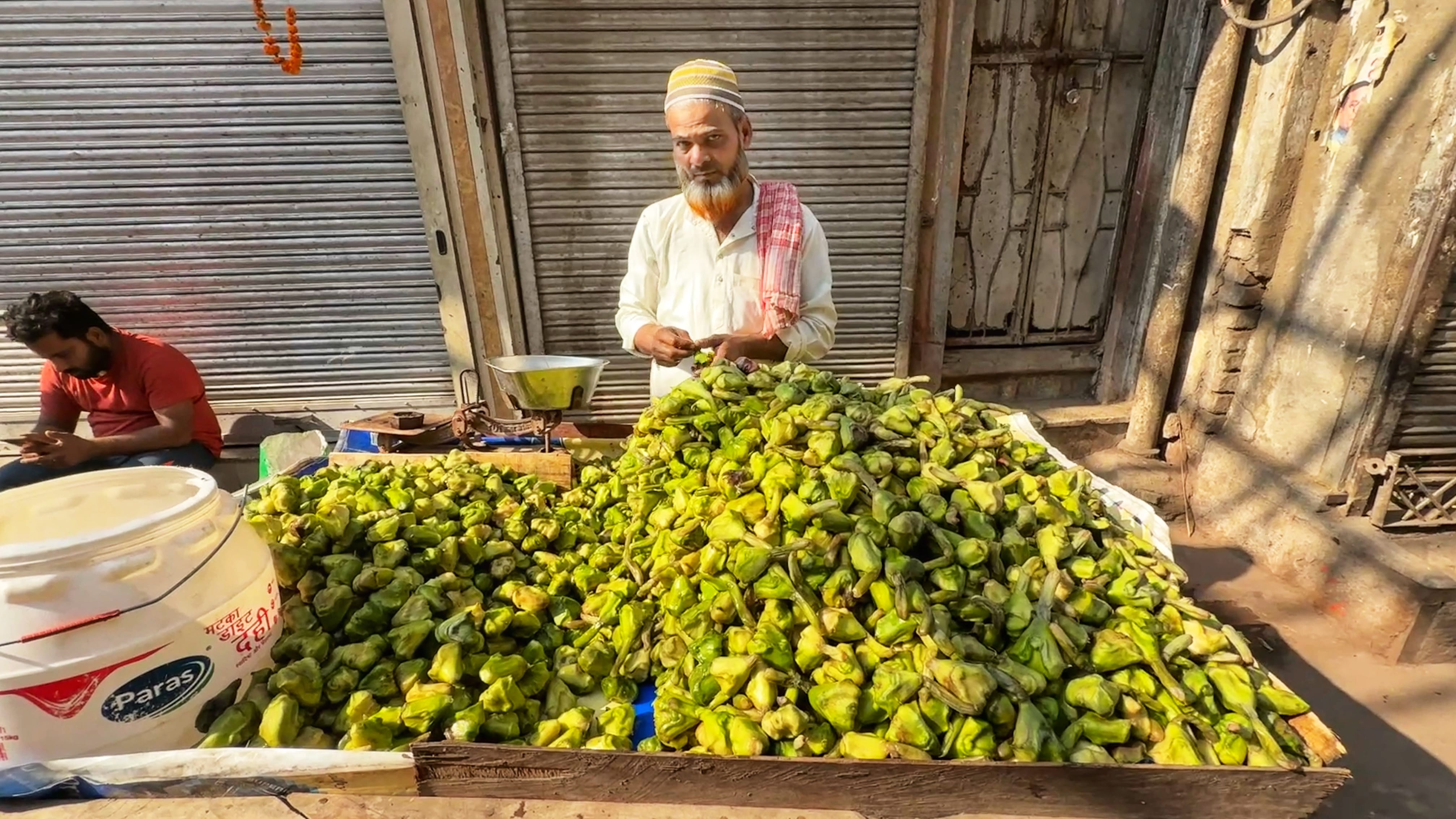 Man selling piles of green singhara (water chestnuts) on a wooden cart in Old Delhi, sitting in front of closed metal shutters
