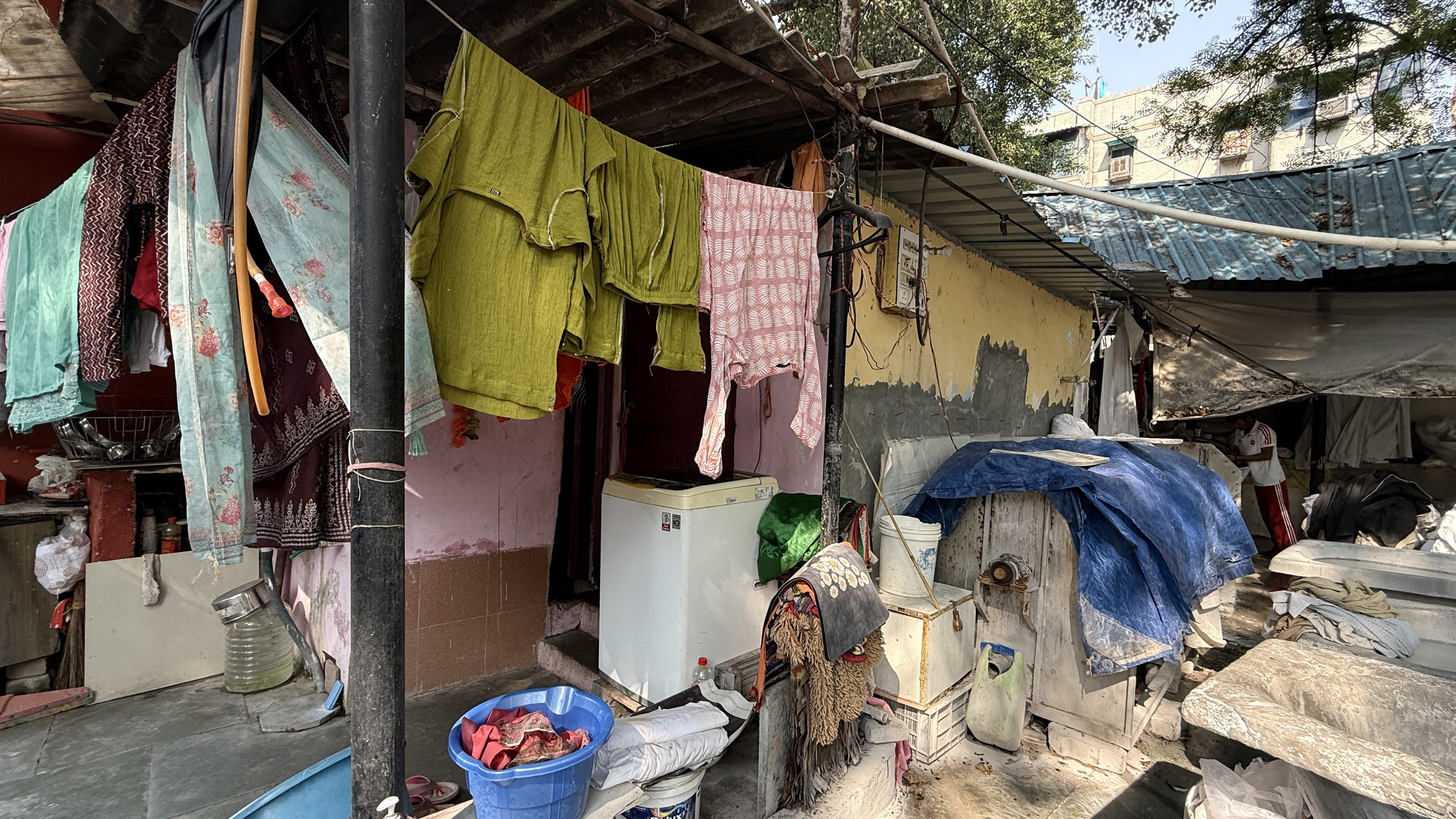 Colorful clothing—greens, red, pink—hanging to dry at a dhobi ghat in Delhi alongside household laundry machines and buckets.