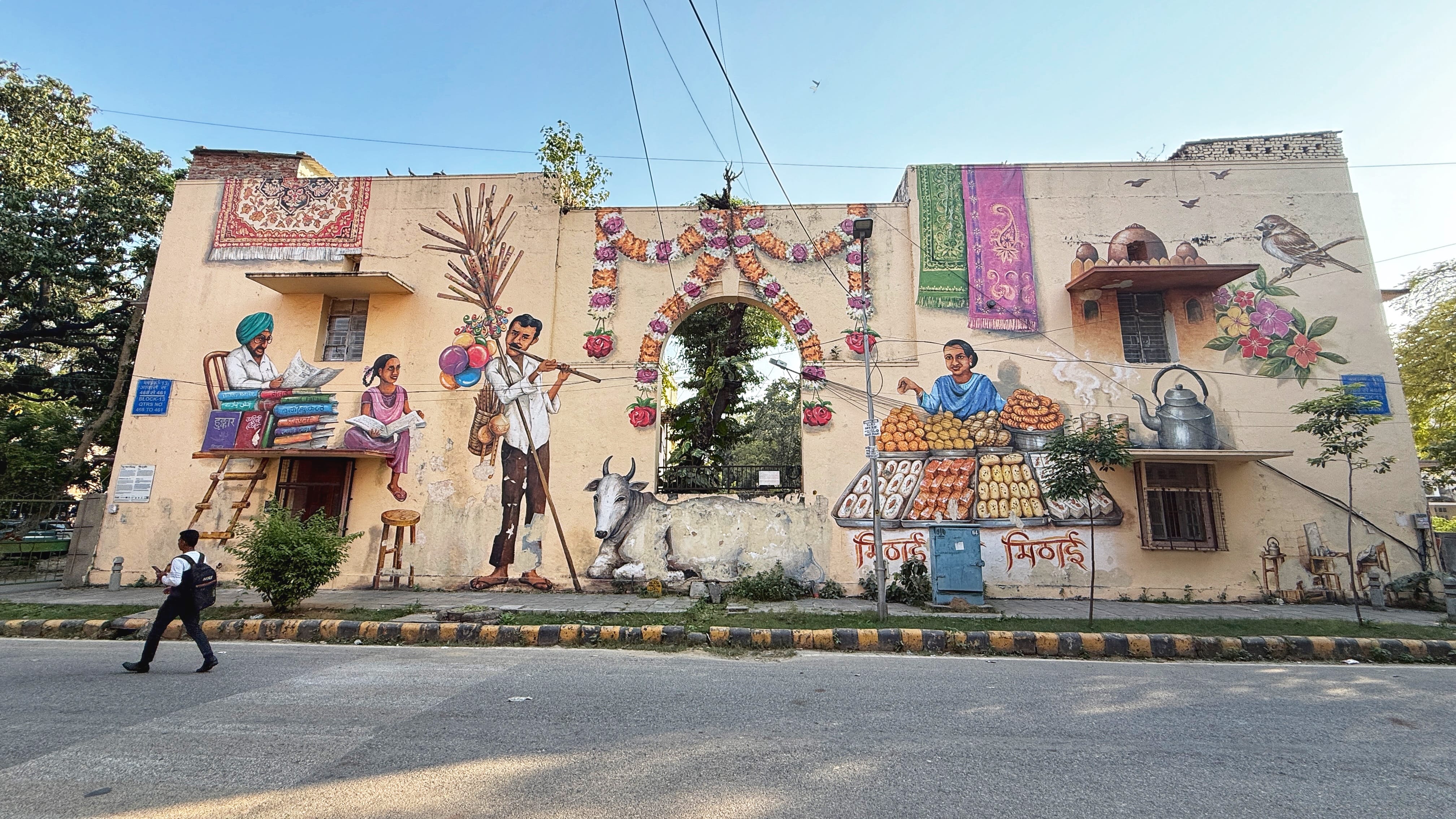 Street art mural at Lodhi Art District in Delhi showing everyday life scenes—vendors selling sweets, a family reading, a cow resting, garlands, textiles, and floral details—2 Days in Delhi
