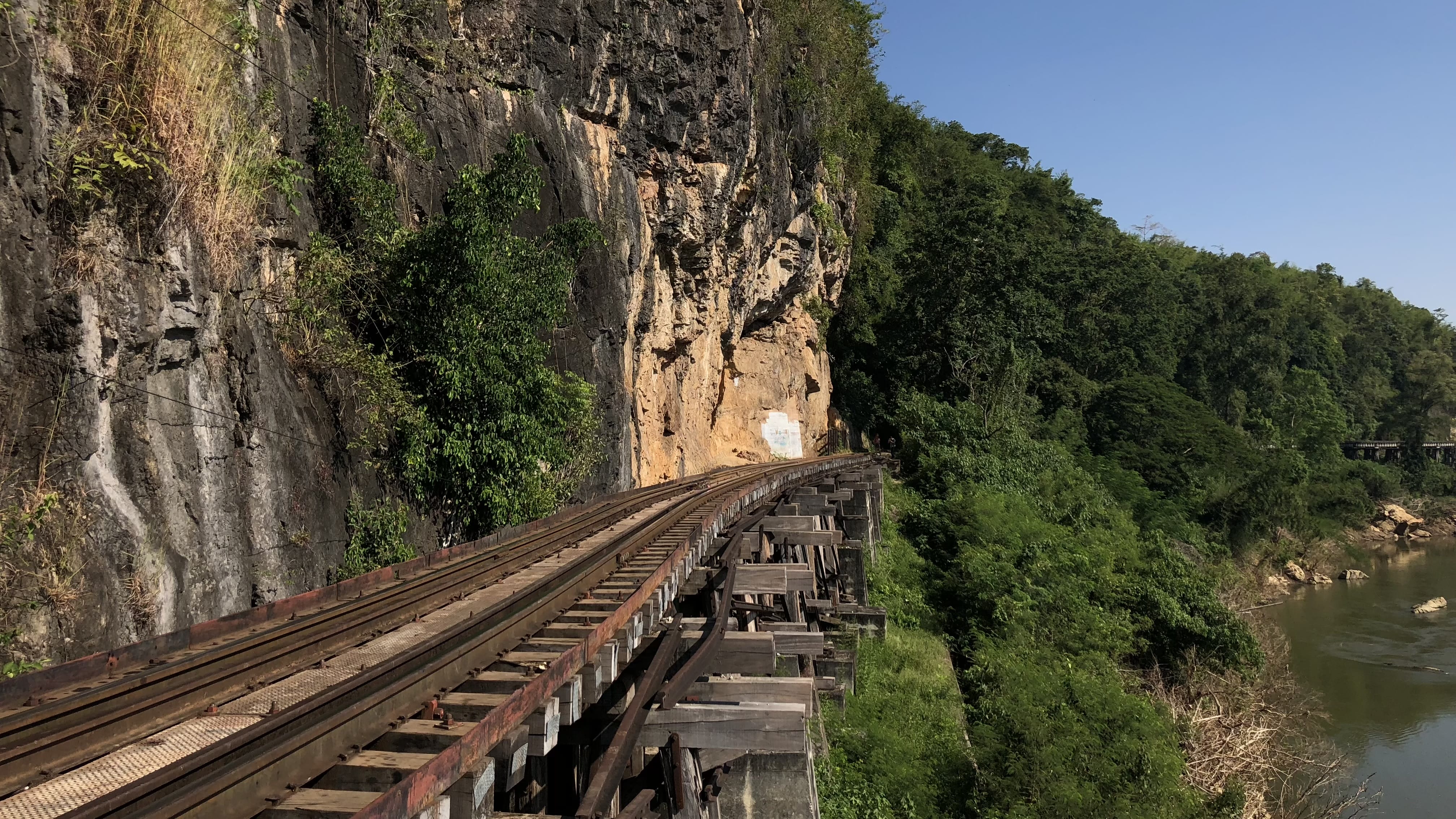 Railway tracks clinging to a cliff along the Thai-Burma Railway near Hellfire Pass, carved into rock and surrounded by dense jungle.