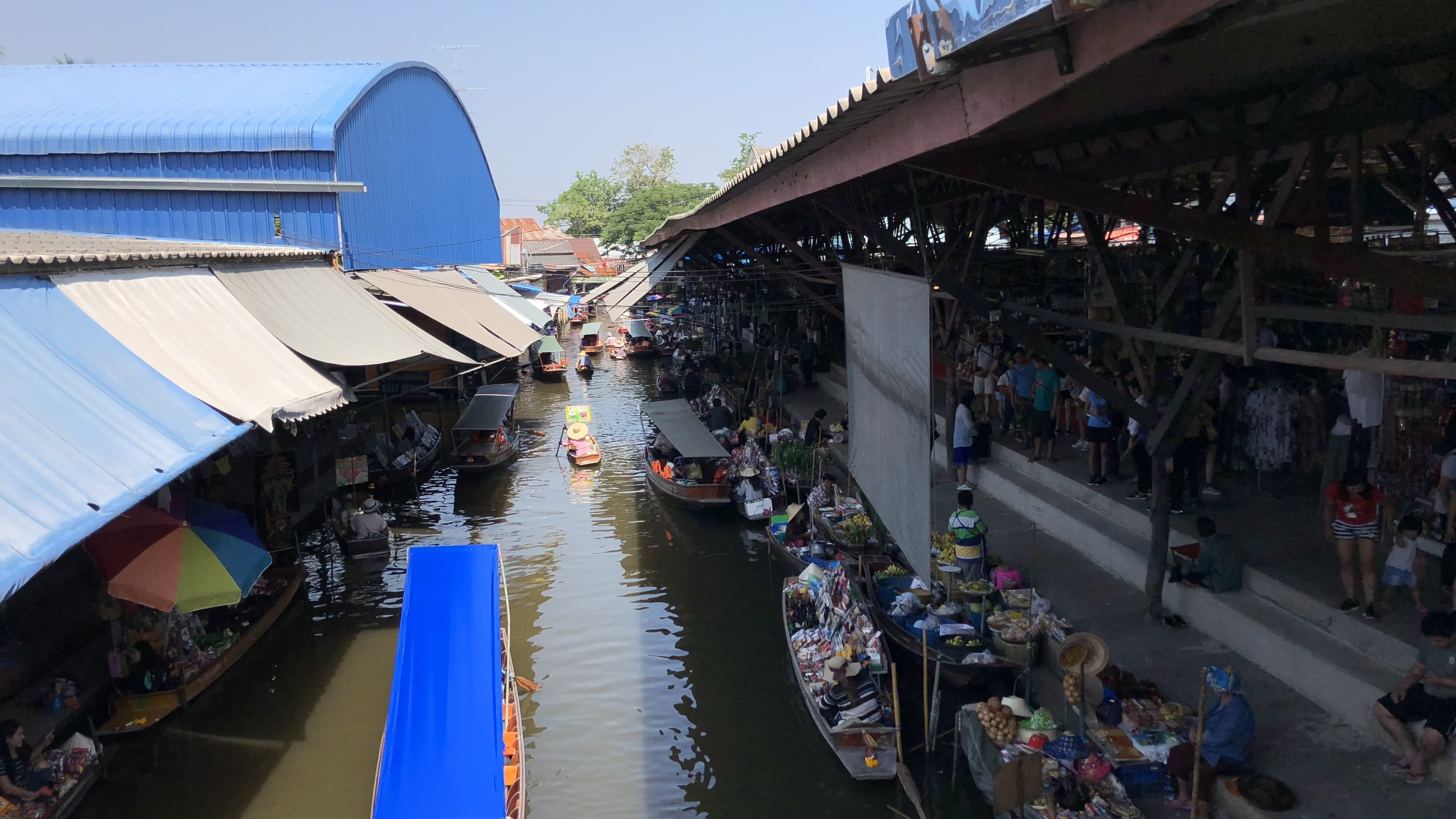 Damnoen Saduak Floating Market with wooden boats packed with produce and goods navigating a narrow canal lined with market stalls in Thailand.