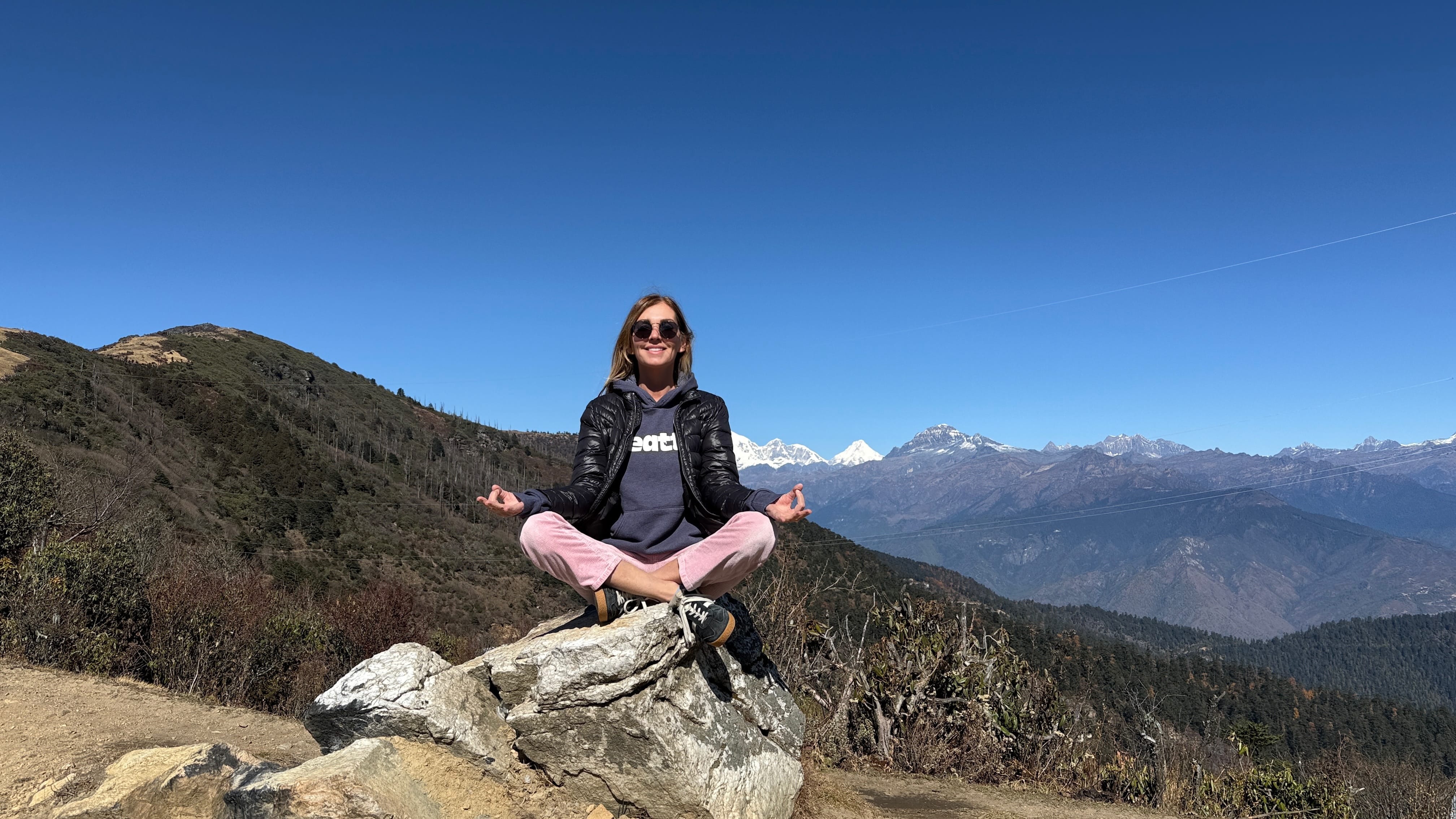 Woman sitting cross-legged on a rock at Chelela Pass in Bhutan, with snow-capped Himalayan peaks and Mount Jomolhari visible in the distance.