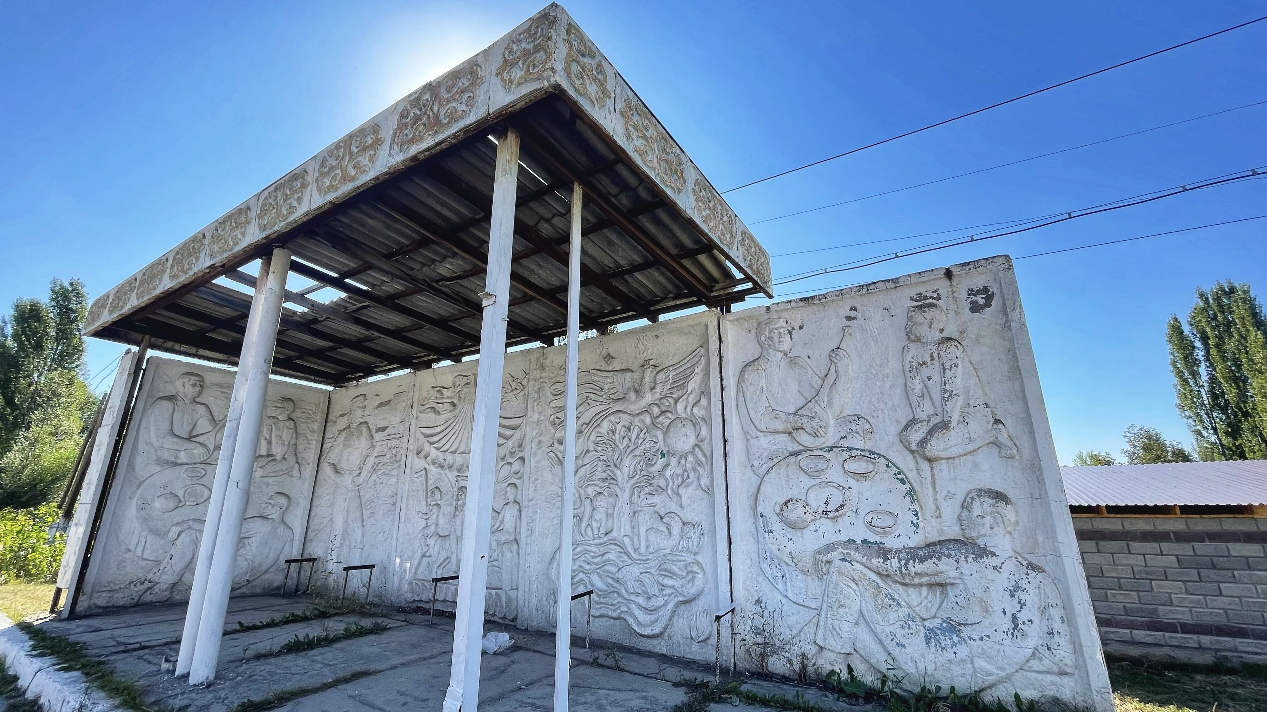 Soviet-era bus stop in Kyrgyzstan with concrete relief panels depicting figures and symbolic scenes beneath a simple metal roof along a rural roadside.