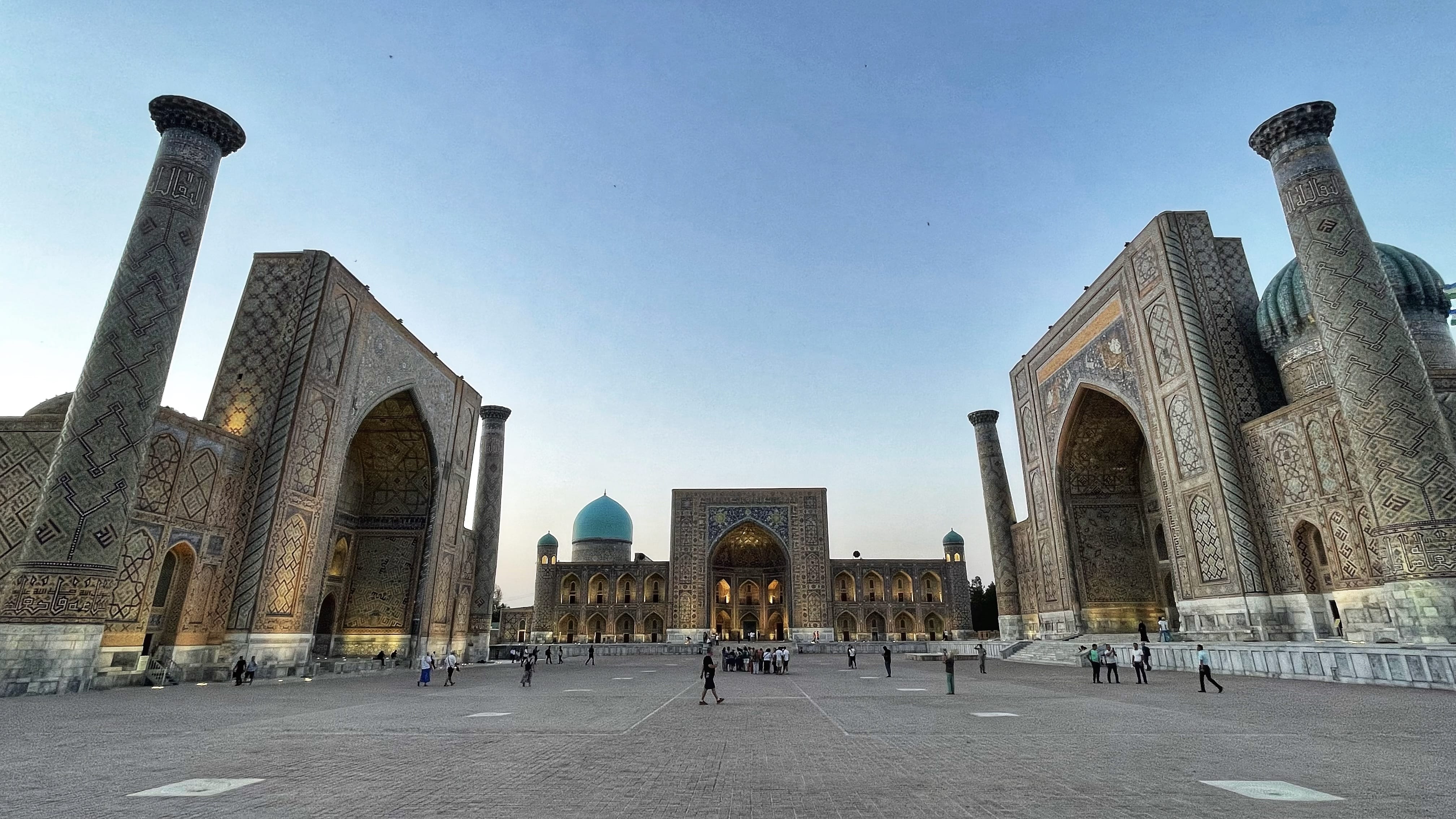 Wide view of Registan Square in Samarkand, Uzbekistan, showing three historic tiled madrasas with towering arches, minarets, turquoise domes, and intricate mosaic patterns at dusk
