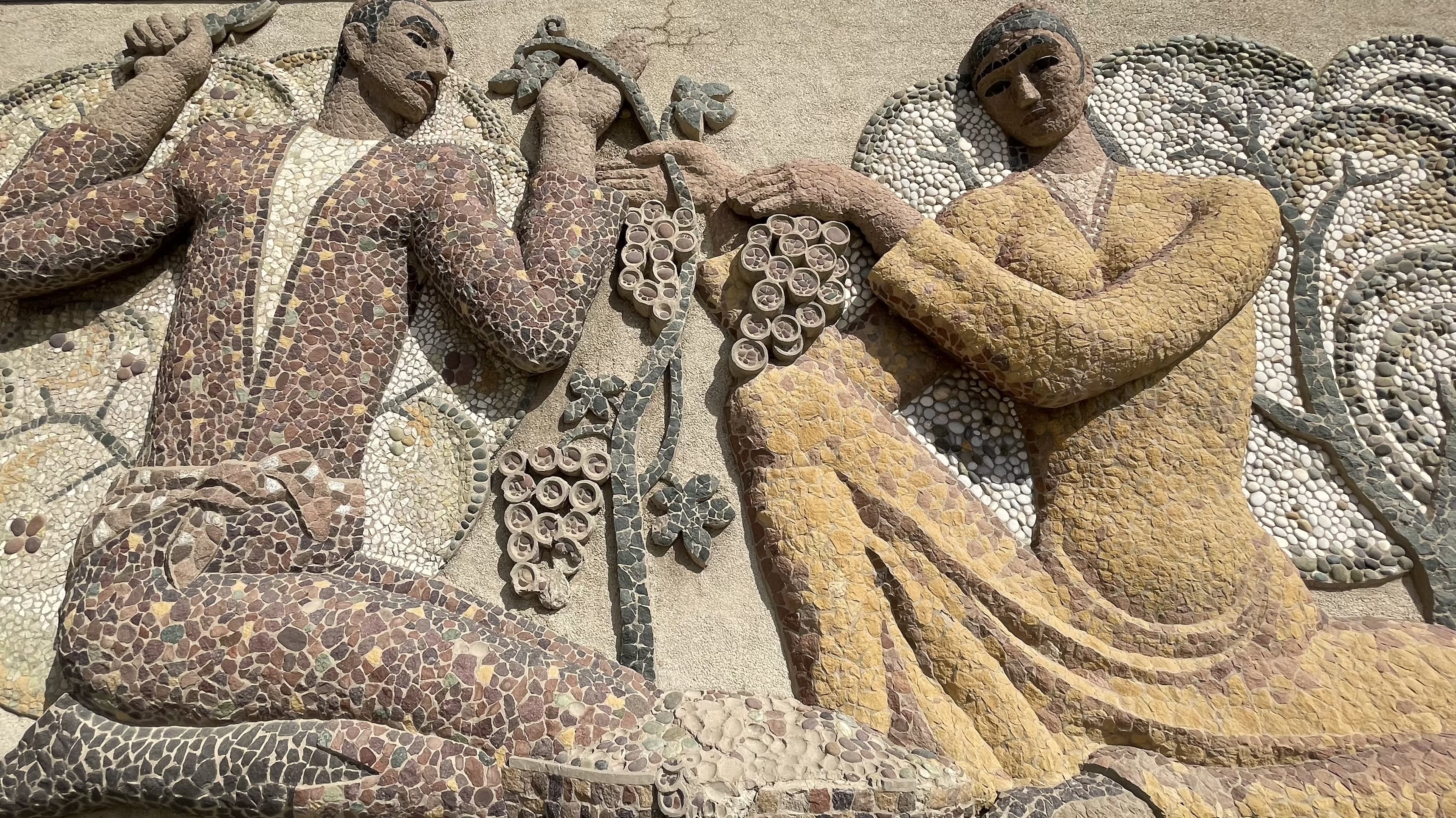 Soviet-era mosaic relief in Tajikistan showing stylized figures holding grape clusters, built in raised stone and tile with deeply textured, sculptural details during our 3 days in Khujand, Tajikistan