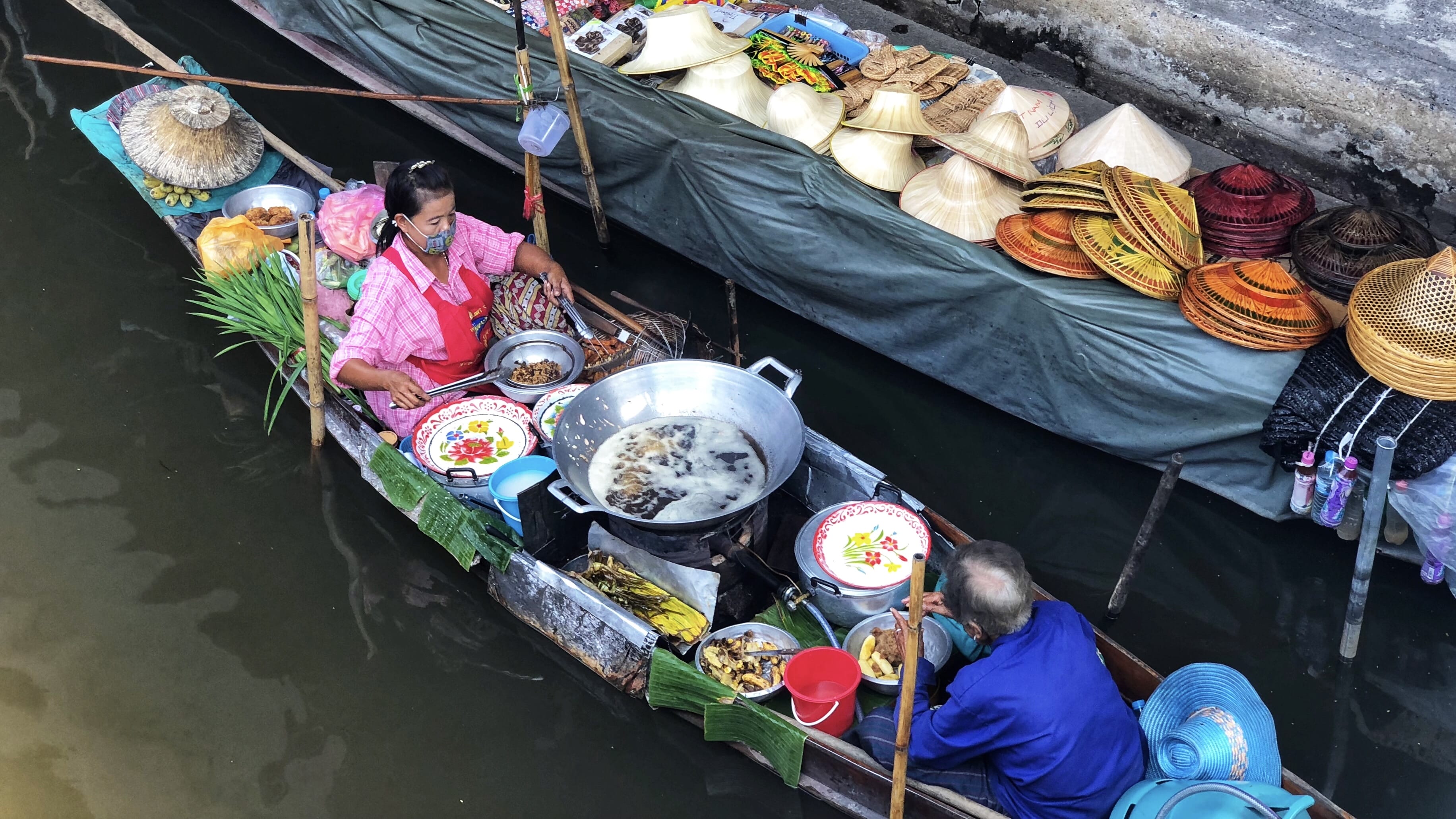 Floating food vendor cooking from a wooden boat at Damnoen Saduak Floating Market, with a large pan of hot oil, ingredients laid out on the boat, and woven hats stacked along the canal edge.