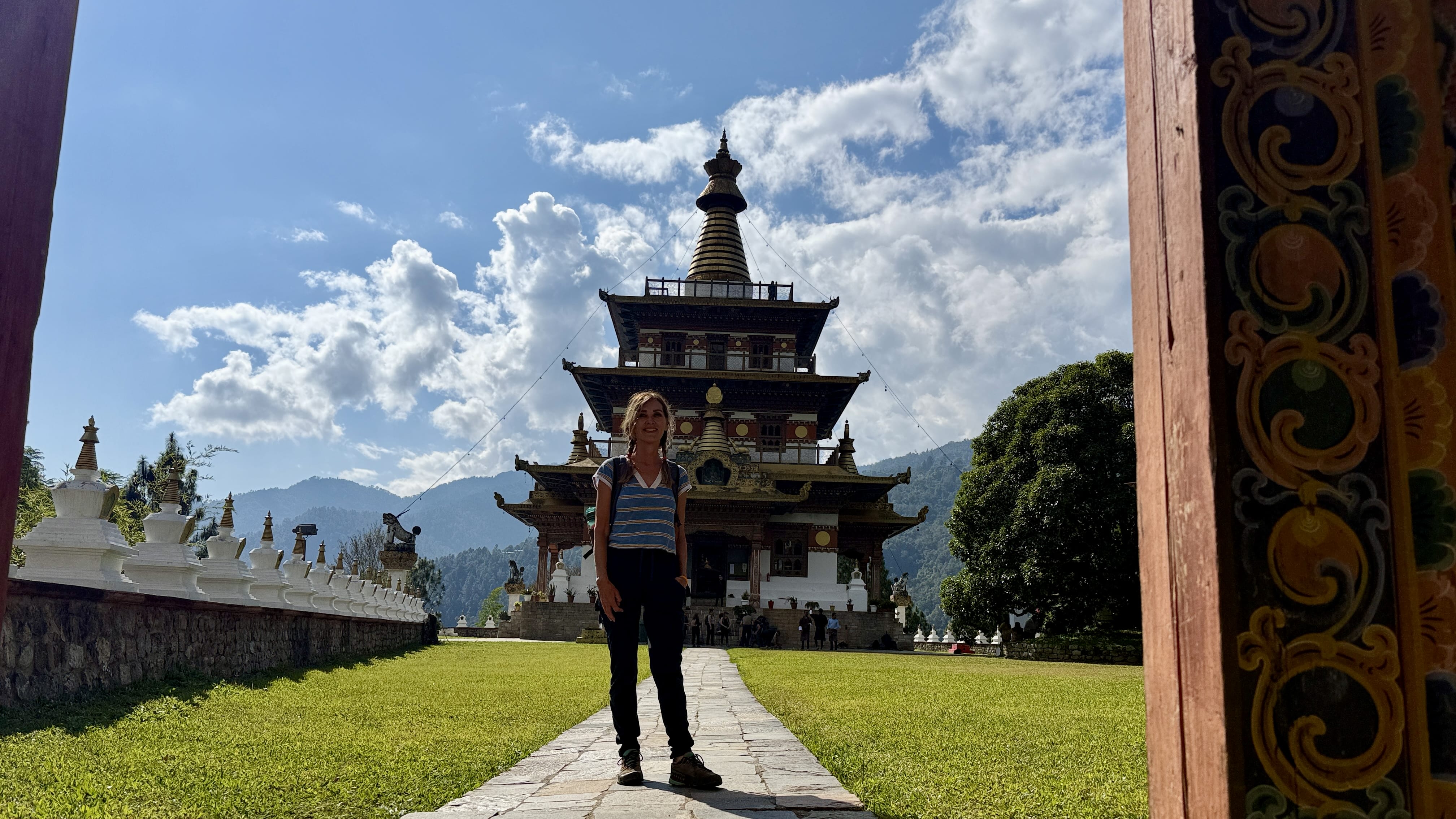 Traveler standing on the stone path at Khamsum Yulley Namgyal Chorten in Punakha Valley, Bhutan, with the multi-tiered chorten and surrounding mountains behind.
