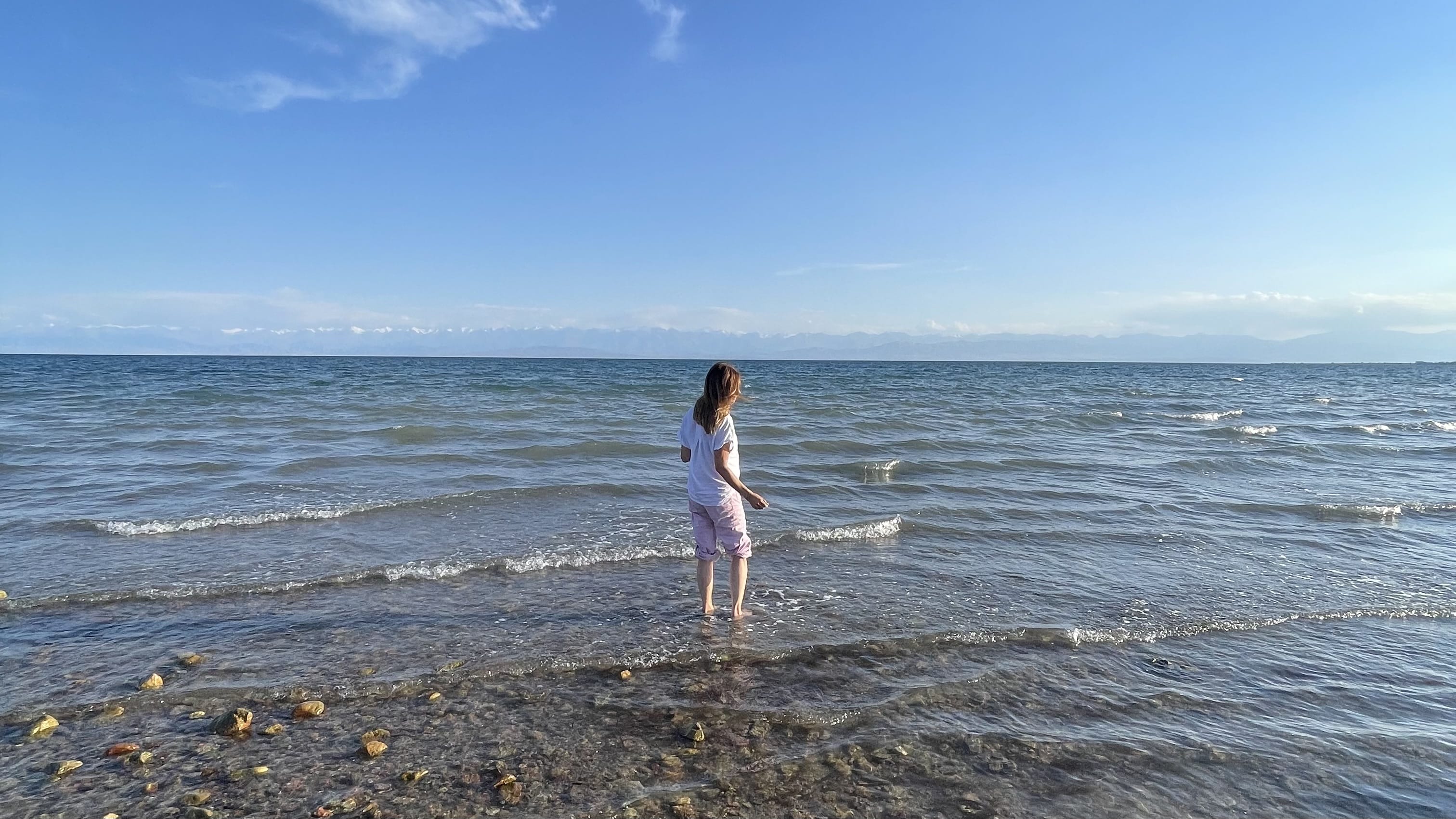 A woman standing barefoot in the clear shallows of Issyk-Kul Lake, with gentle waves and distant mountains on the horizon
