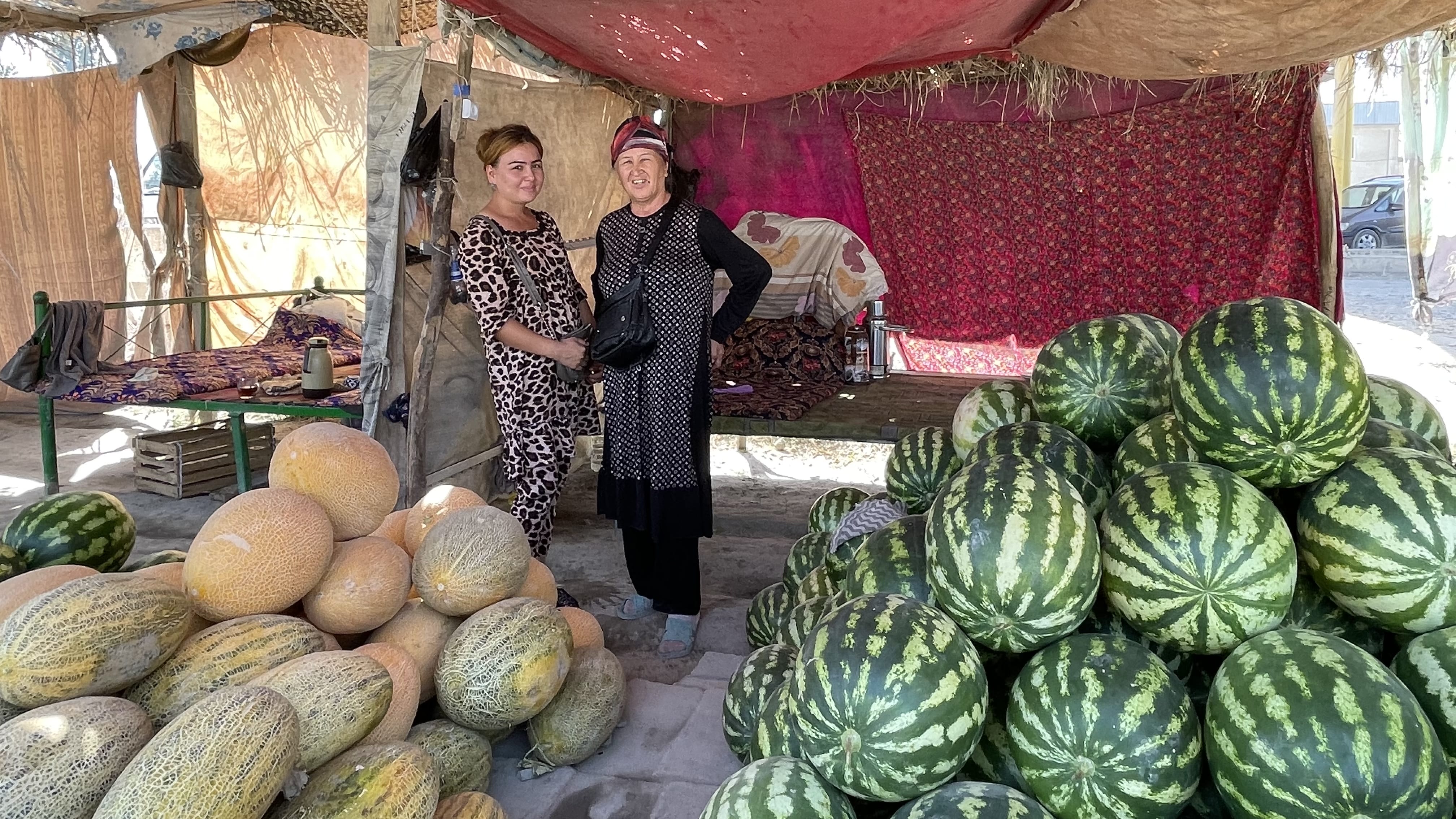 Roadside fruit stand near Khujand, Tajikistan, with piles of watermelons and melons and two women standing under a shaded canopy, photographed during a 3 days in Khujand, Tajikistan itinerary.