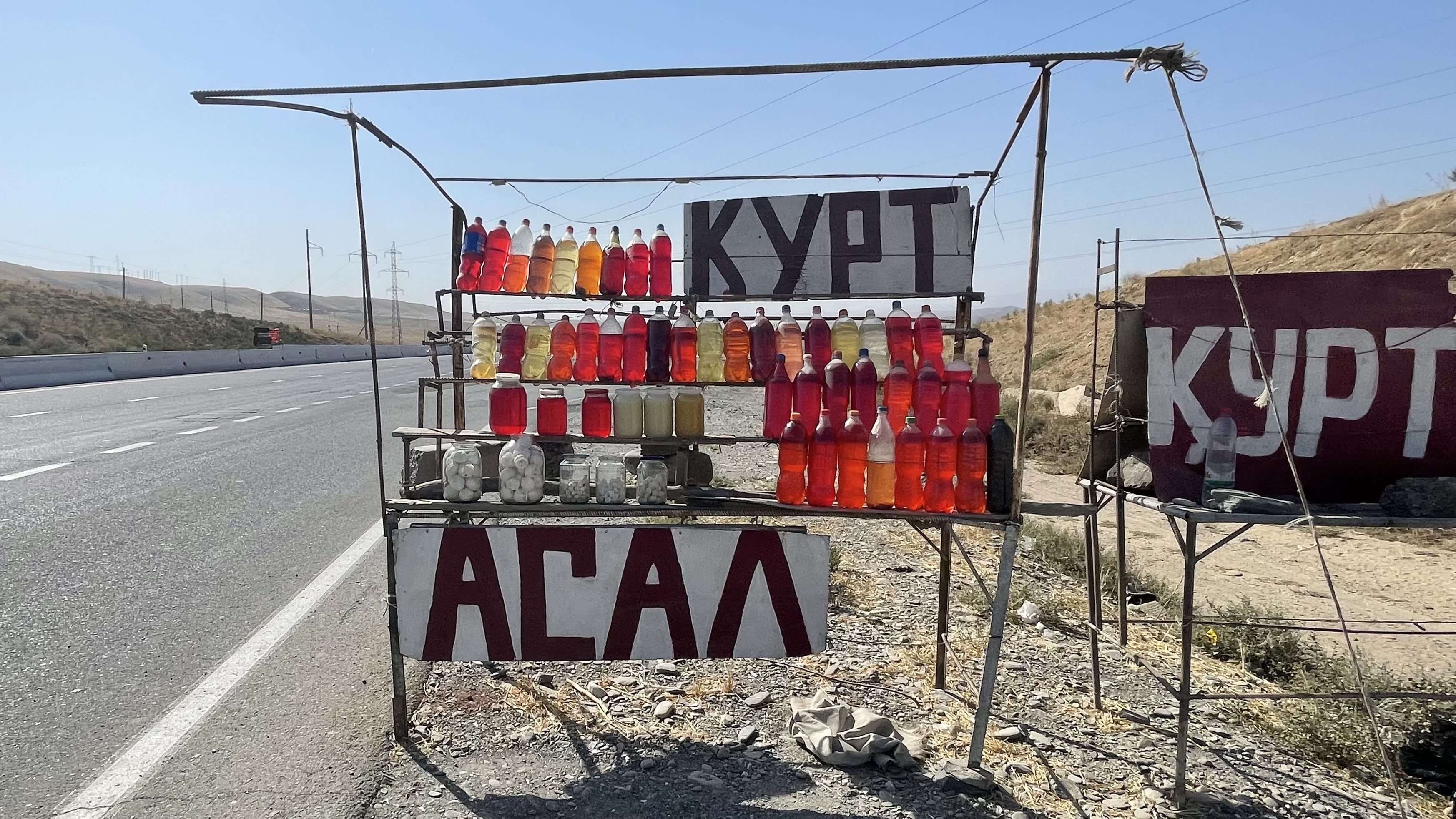 Roadside fuel stand in Uzbekistan selling gasoline by the liter in reused soda bottles, with jars of mothballs displayed beside the bottles along a rural highway
