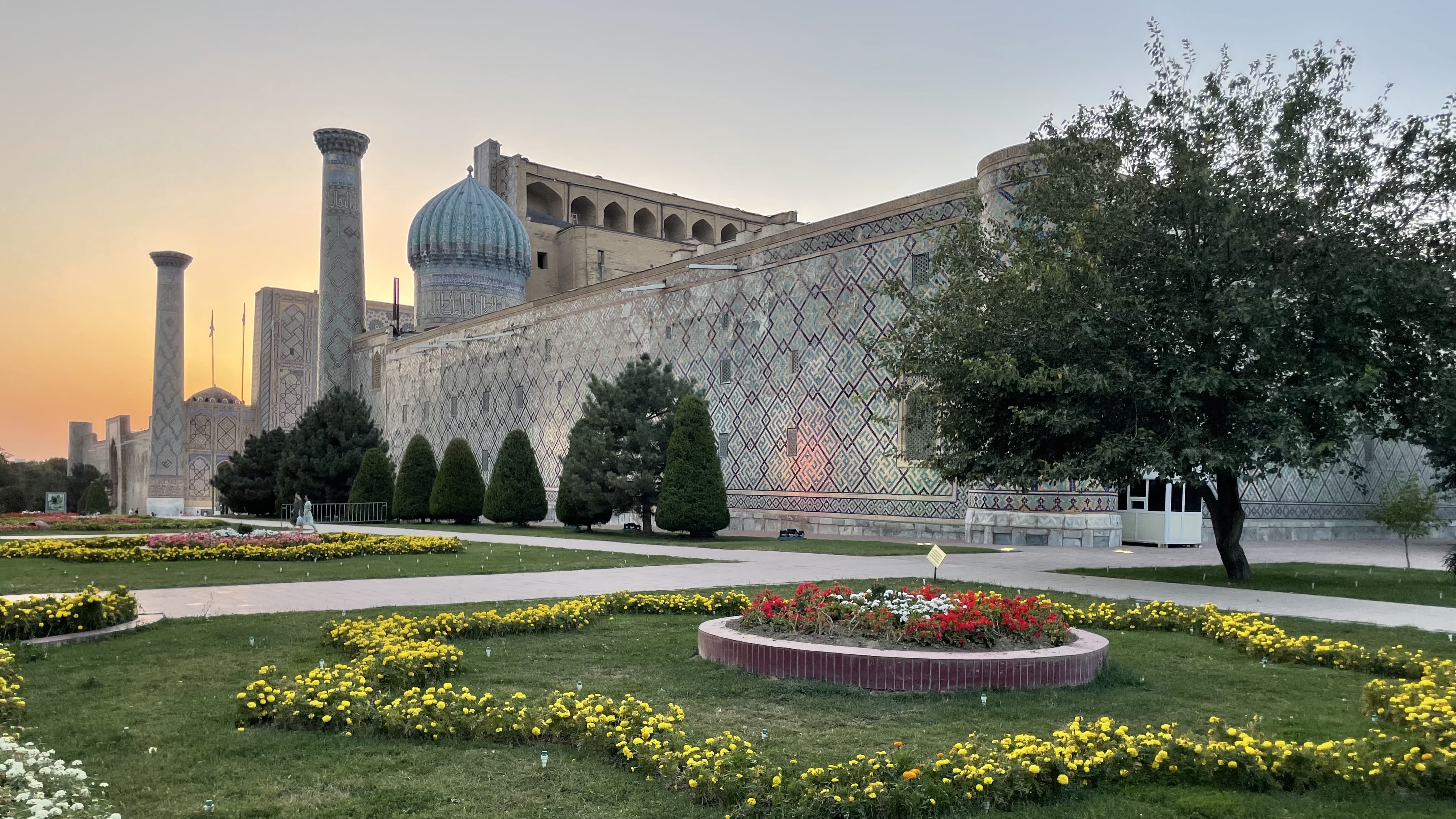 Registan Square in Samarkand at sunset, showing tiled madrasas with turquoise domes, patterned mosaic walls, and formal gardens in Uzbekistan