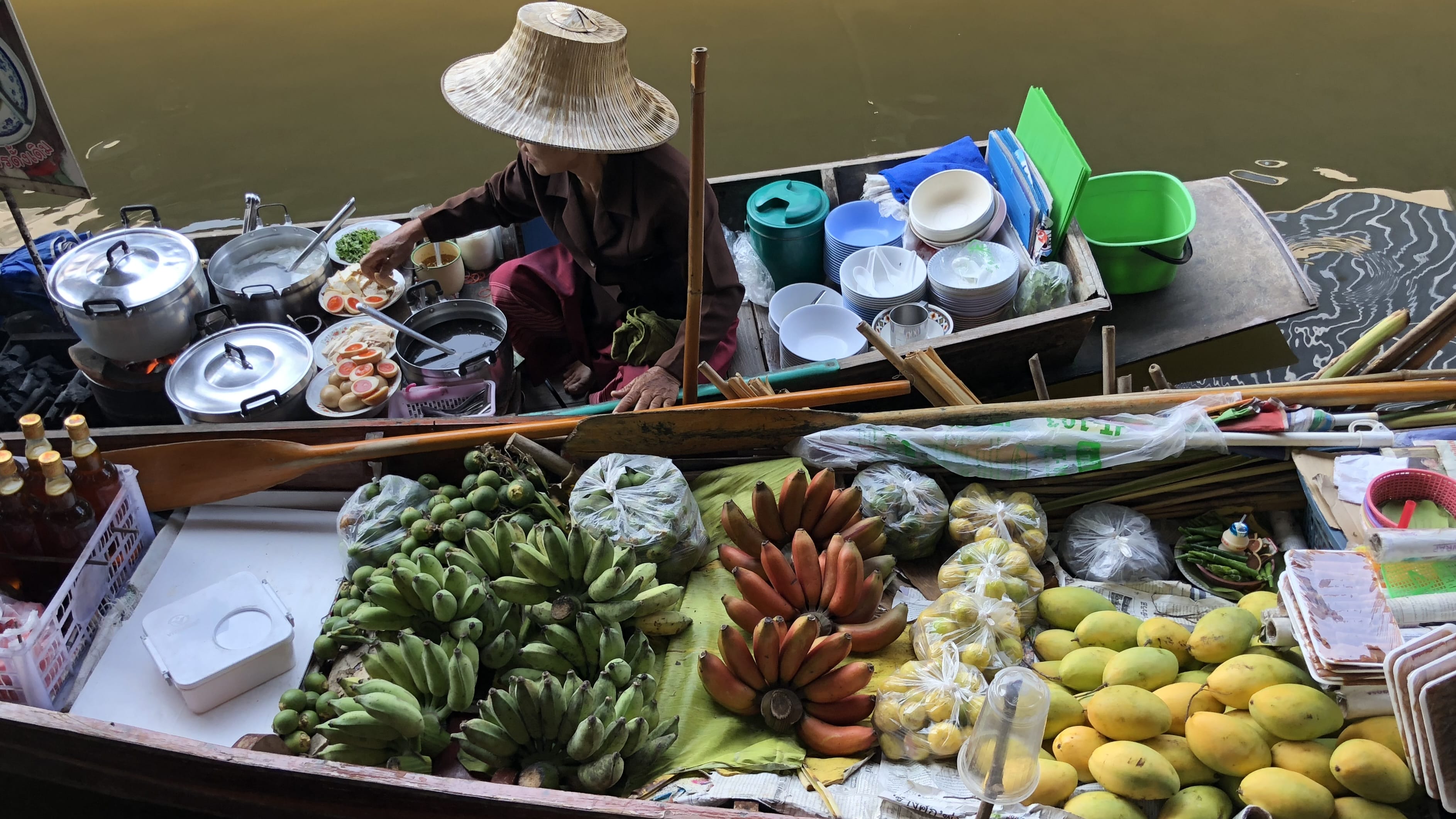Floating food vendor preparing dishes from a wooden boat at Damnoen Saduak Floating Market, with pots, bowls, sliced eggs, and baskets of bananas and mangoes arranged along the canal.
