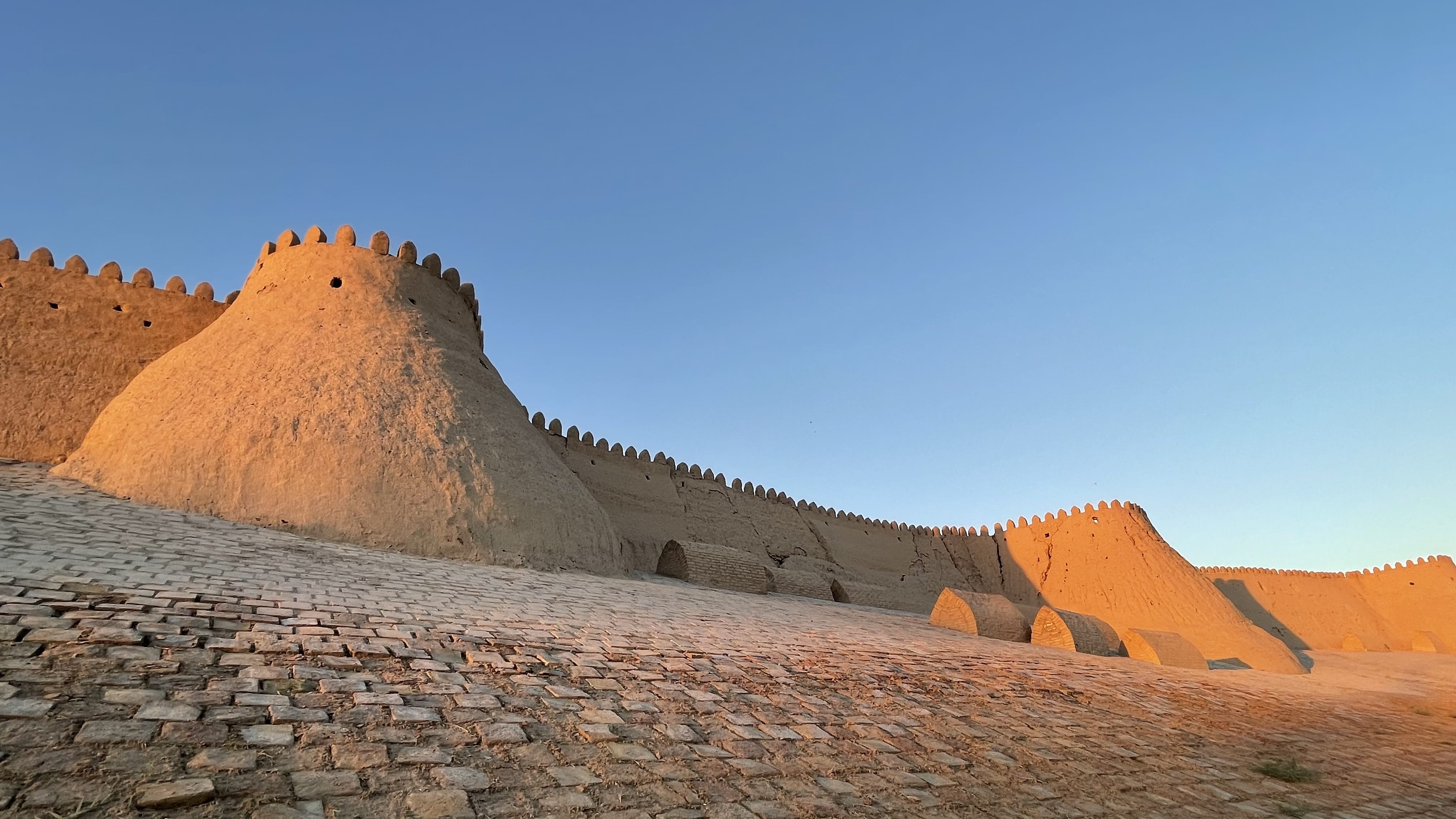 Alt text (SEO): Sunset light on the mud-brick fortress walls and rounded bastions of Itchan Kala (Ichan-Qala) in Khiva, Uzbekistan, with a cobblestone slope in the foreground and clear blue sky above.