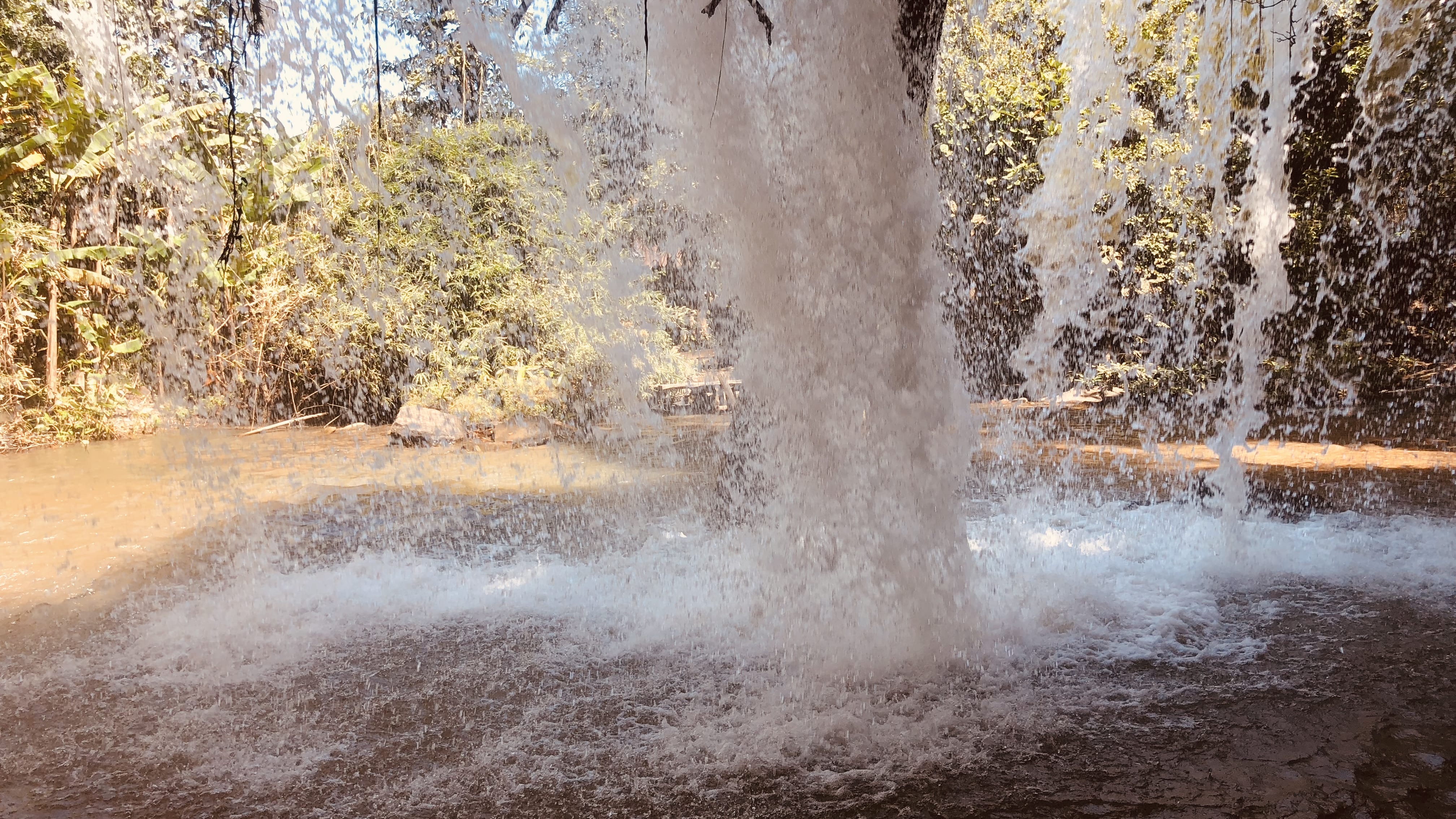 Water cascading down a wide waterfall on the Mae Wang Waterfall Trail in northern Thailand, viewed from behind the falling water with a shallow pool and forest beyond
